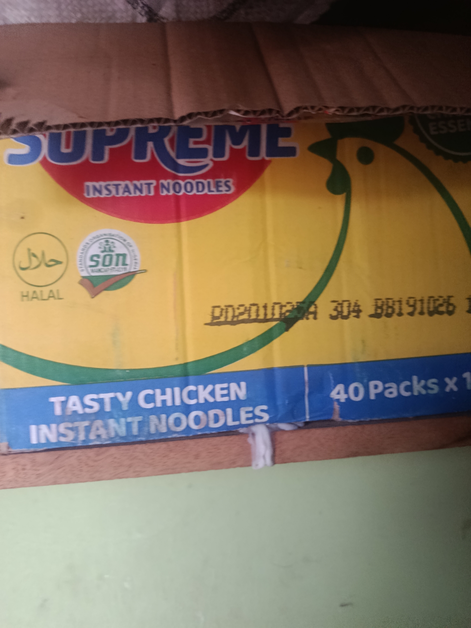 The image shows a yellow cardboard box with blue and red text. The text "SUPREME" is prominently displayed in large blue letters at the top of the box. Below it, in smaller red letters, is "INSTANT NOODLES". To the left of the "INSTANT NOODLES" text, there are two circular logos. The left logo is green and contains Arabic text that appears to say "Halal". The right logo is circular and white with green accents, featuring a stylized image of a chicken and text that includes "SON" and "NIGERIA". To the right of these logos, a portion of a yellow chicken illustration is visible. A black printed serial number or code, "D020102AA 304 BB191026 1", is also visible on the yellow surface.
The bottom portion of the box has a blue band with white text. The text reads "TASTY CHICKEN INSTANT NOODLES" on the left and "40 Packs x 1" on the right. The cardboard box appears to be sitting on a wooden surface, and behind it, a textured grey material is visible, possibly a wall or shelf. The lighting is dim, and the image is slightly blurry, suggesting it was taken in low light conditions. The overall scene suggests a product package, likely for food items. There are no people or discernible actions taking place in the image. The setting is indoors, but no specific city or location is identified.