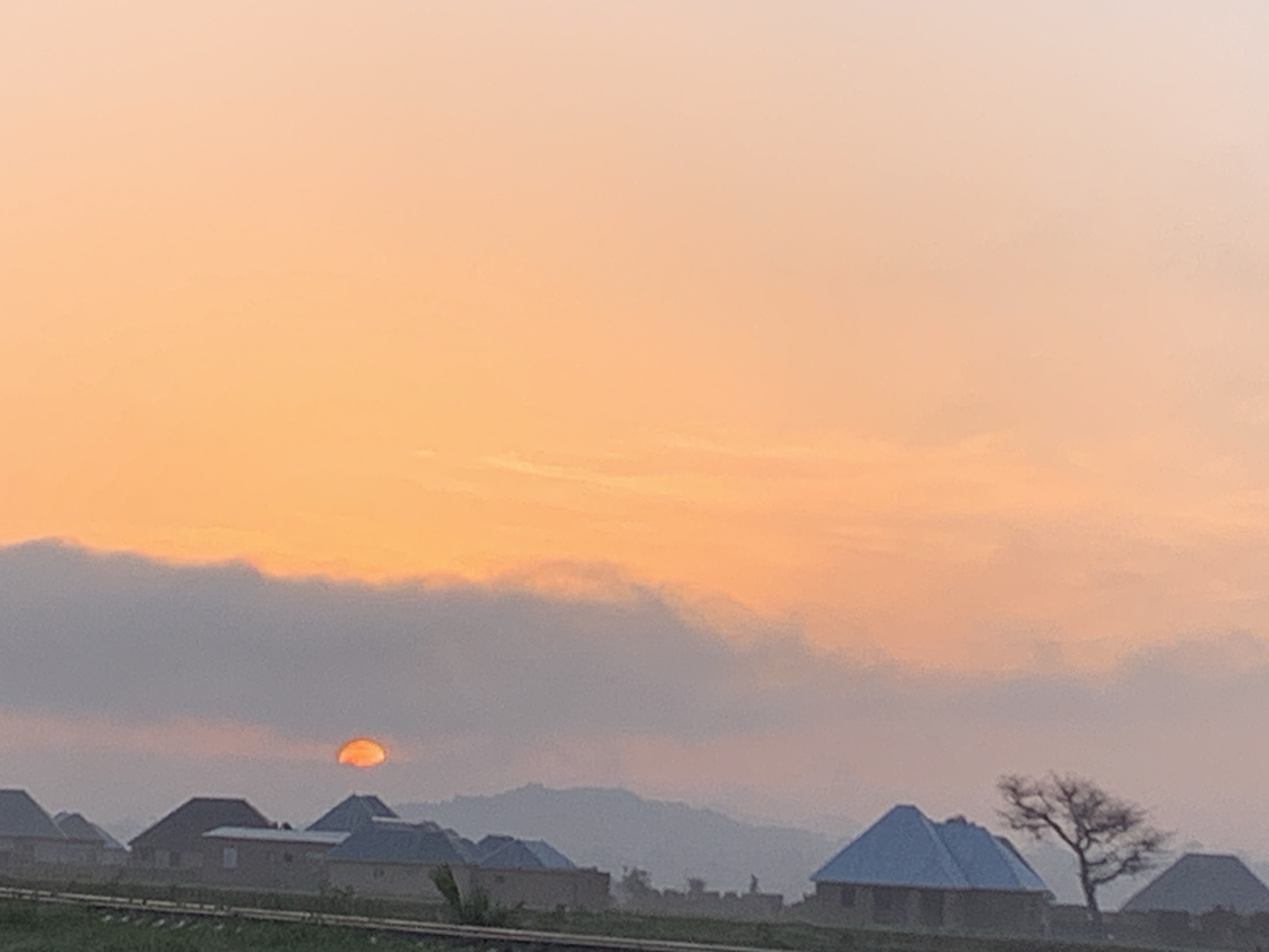 The image depicts a serene dawn scene in Kuru Karama, Nigeria. The sky is dominated by a soft, gradient of orange and peach hues, indicative of a sunrise. A hazy sun, partially obscured by a layer of clouds, casts a warm glow. In the foreground, several houses with blue roofs are visible, nestled amongst lush greenery. A lone, bare tree stands to the right of the houses, silhouetted against the bright sky. In the distance, rolling hills create a gentle backdrop to the settlement. The overall impression is one of tranquility and the quiet beauty of a new day beginning in this Nigerian locale.