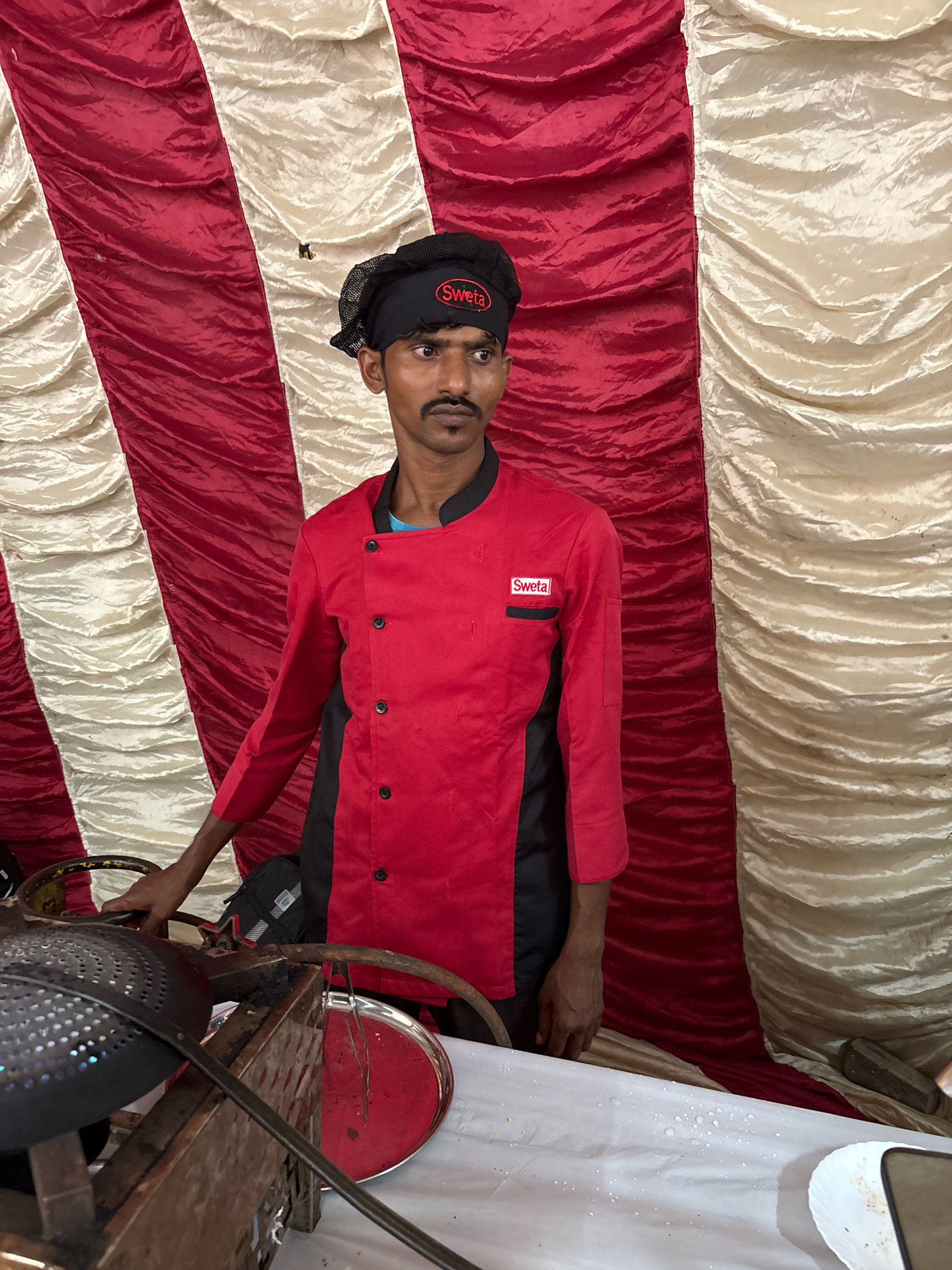 A man wearing a red chef's jacket with a black mesh cap stands in front of a red and cream striped fabric backdrop. The jacket has a black collar and black panels on the sides, with black buttons down the front. A red and white "Sweta" logo is visible on the left breast pocket. He has a mustache and is looking to the right. In the foreground, a metal rack holds cooking equipment, including a black colander with holes and a red plate. The scene appears to be set up for outdoor food preparation or a food stall. The location is specified as Howrah, India.