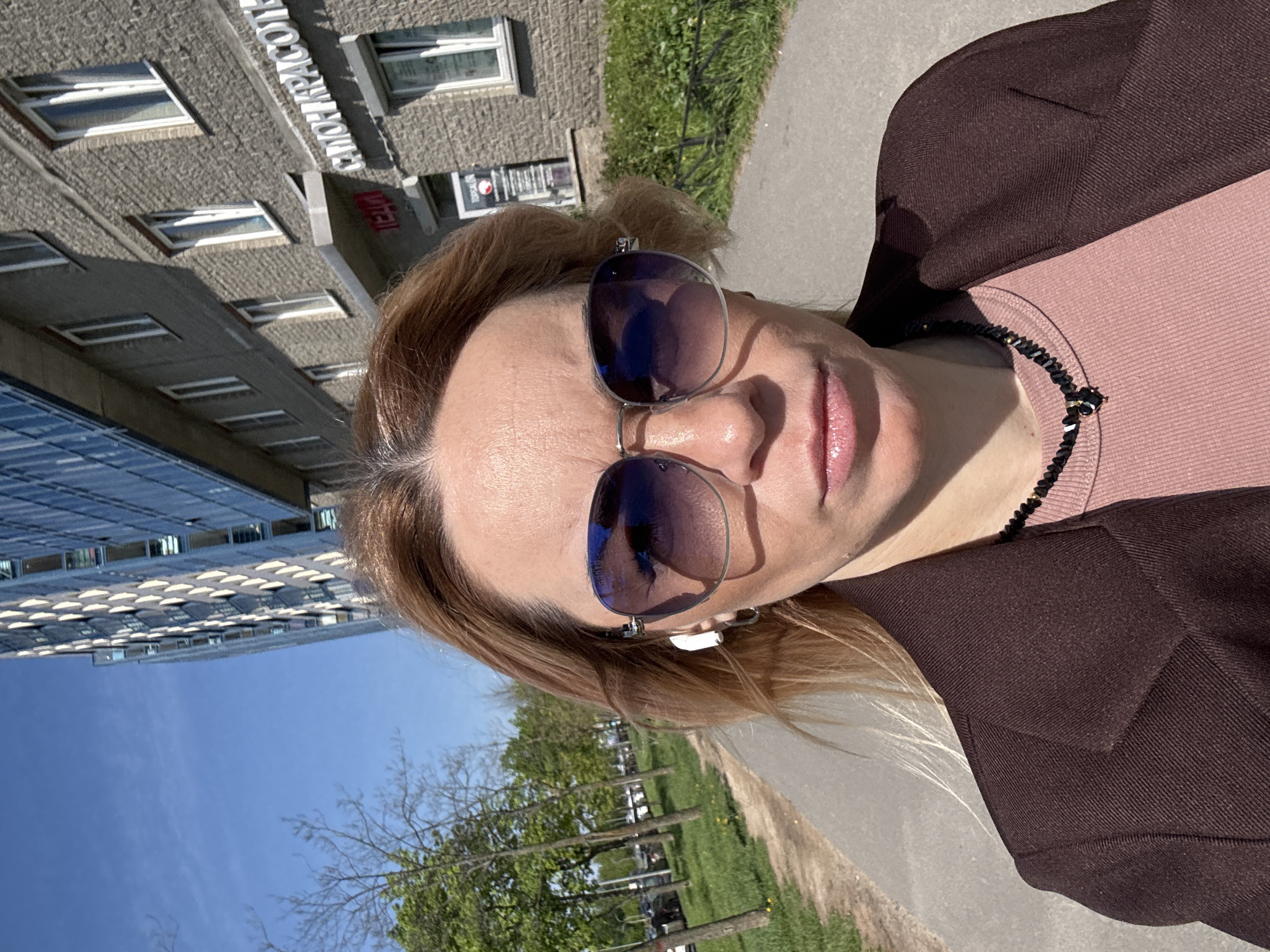 A woman is standing outside, seemingly taking a selfie. She's wearing sunglasses, a pink top, and a brown jacket. She also has what appear to be wireless earbuds in her ear and a beaded necklace. In the background, there's a building, trees, and a blue sky. The building in the background has an awning with the sign in Russian Cyrillic "СИМОН КРАСОТА" which translates to "Simon Beauty". The scene is located in Vsevolozhsk, Russia, based on the Cyrillic sign.