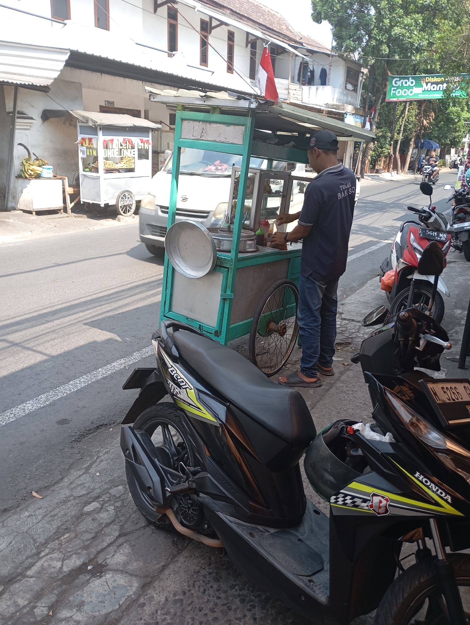 The image depicts a street scene in Malang, Indonesia, likely a commercial area given the presence of food vendors and parked motorcycles. In the foreground, two black motorcycles are parked on the side of the road. One is a Honda Beat, recognizable by its design and branding, and the other is partially visible. A man wearing a dark t-shirt with "TULUS BAKSO" printed on the back and blue jeans is standing at a food cart. The cart itself is green and white, with a display of food items and various bottles of condiments. In the background, there is a white building with multiple windows, a red and white Indonesian flag, and trees lining the street. A "Grab Food Mantap" sign is also visible, indicating a food delivery service is available. Another food cart selling what appears to be bananas is situated to the left of the main food cart. The road surface shows signs of wear and tear, and shadows are cast across the scene, suggesting it is daytime.