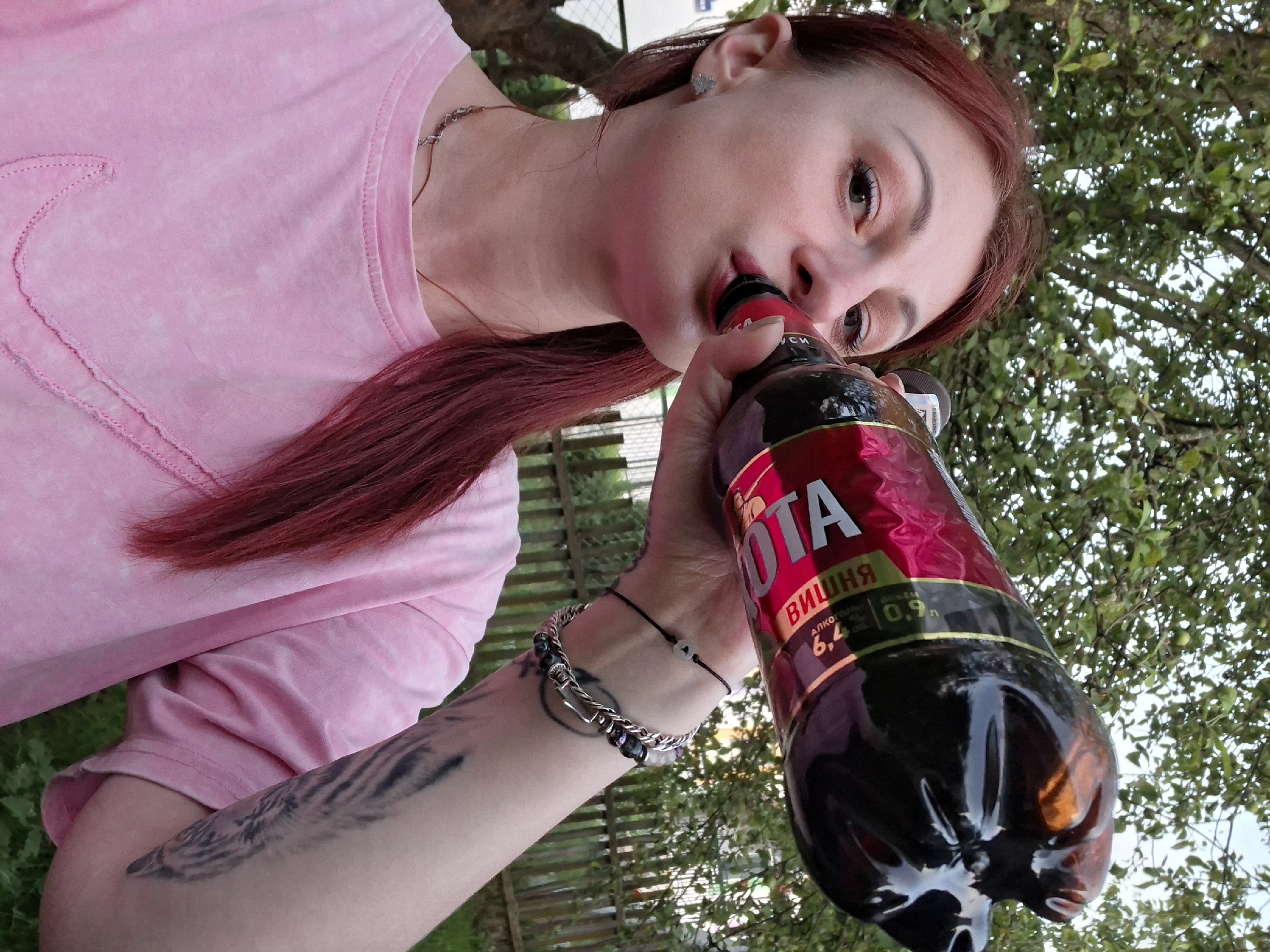 In this eye-level, close-up shot, a young woman with long, reddish-brown hair is drinking from a large, dark-colored plastic bottle of beverage. She is wearing a pink t-shirt and a silver necklace. Her left arm, which is holding the bottle, is adorned with a tattoo and a bracelet. The setting appears to be outdoors, with a wooden fence and green foliage visible in the background. The lighting suggests it is daytime, possibly late afternoon. The woman's facial expression is neutral as she drinks.
Visible text on the bottle includes "OTA" in large silver letters and "ВИШНЯ" (which translates to "cherry" in Russian) in smaller text below it. Further down, there is text indicating "алкоголь" (alcohol) followed by "6,4 % об." (6.4% by volume) and "0,9 л" (0.9 liters). The context provided places this scene in Забалоцкі сельскі Савет, Belarus.