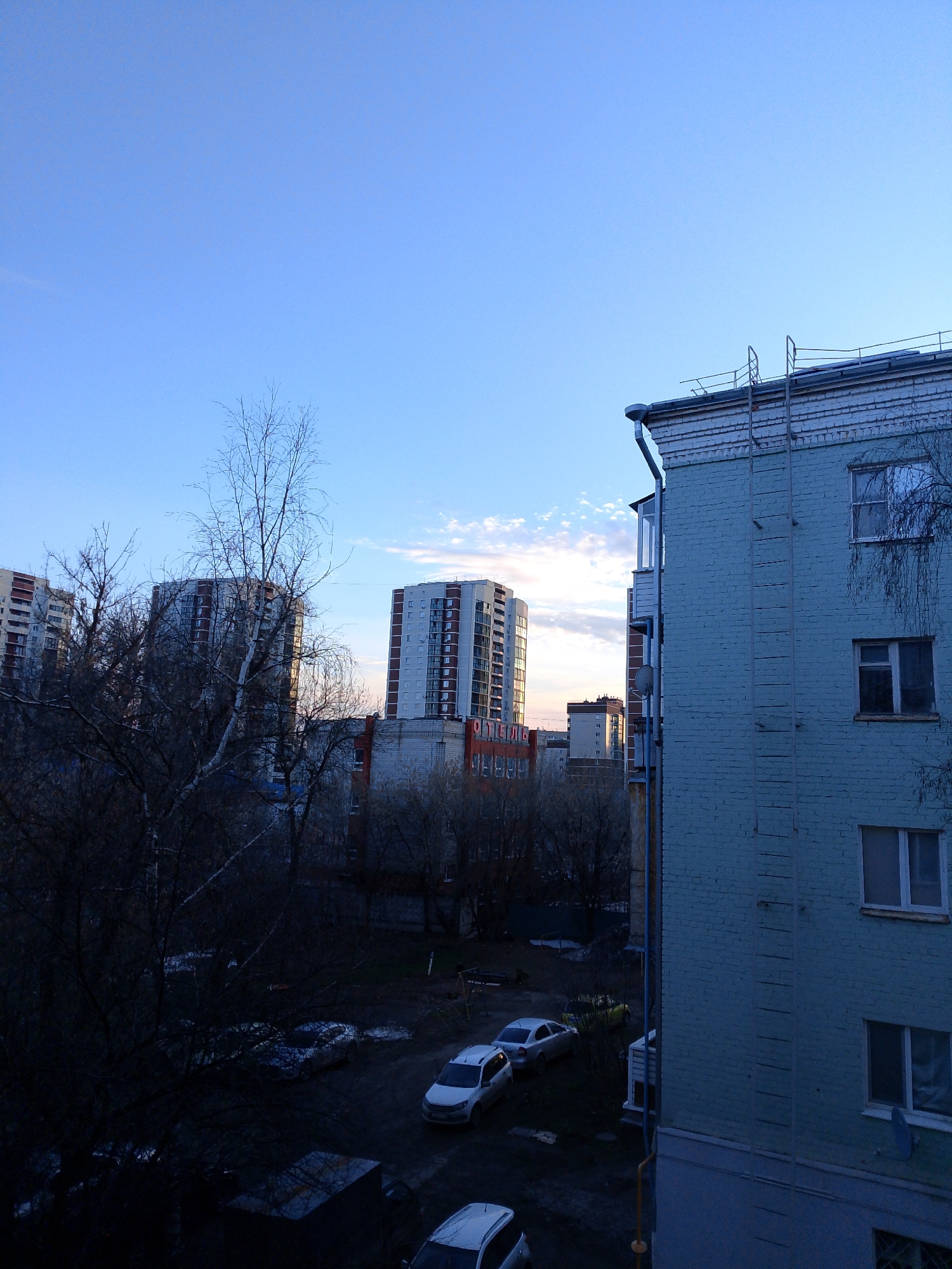 The image shows a view from a high vantage point in Izhevsk, Russia, capturing a residential area at what appears to be dusk. In the foreground is a light-blue brick building with a fire escape ladder attached to its exterior wall. Adjacent to the building, a view opens up to a courtyard with several parked cars on a muddy surface. Beyond the courtyard are multi-story apartment buildings, with the fading sunlight hitting their upper portions. The sky is a gradient of blue, transitioning to orange near the horizon, suggesting either sunrise or sunset. Bare trees stand between the vantage point and the distant buildings.