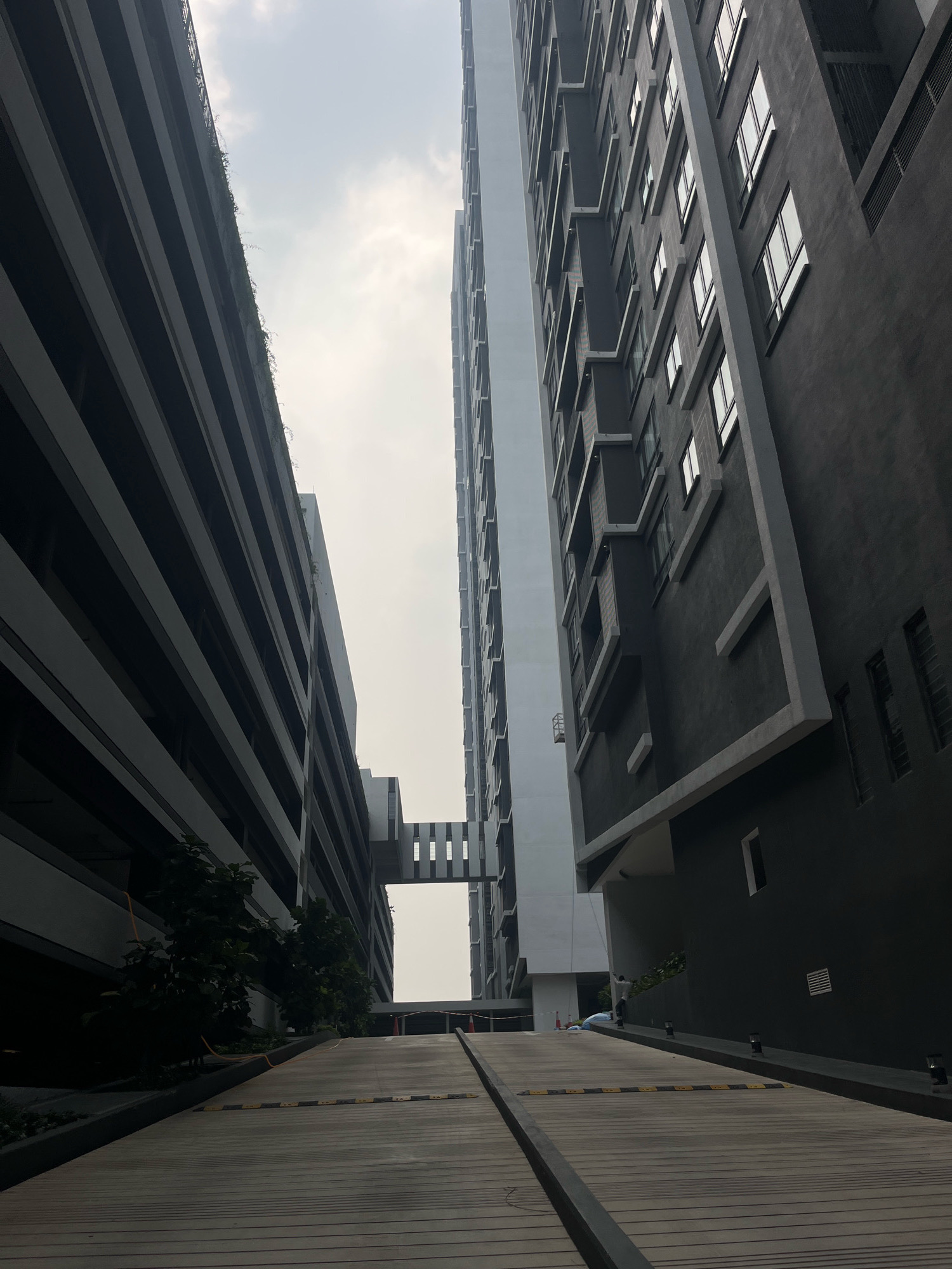The image depicts a modern multi-story building complex, likely an apartment building or condominium, with a concrete ramp leading upwards. The architecture features clean lines and a contemporary design, with the buildings having dark gray and white facades. One building on the right has numerous windows evenly spaced across its surface, while the building on the left appears to be a parking structure with horizontal banding. A skybridge connects the two structures in the distance. The scene suggests an urban environment within Kuala Lumpur, Malaysia, given the architectural style common in the city. The sky is overcast, indicating a cloudy day. There is a lone figure visible in the distance, standing on the ramp. The ramp itself is made of wood-like planks and has speed bumps.