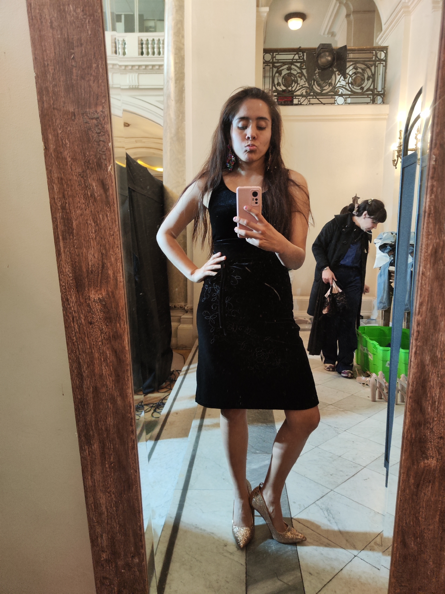 A woman is taking a mirror selfie in a hallway with marbled floors. She is wearing a black sleeveless dress with a floral pattern, sparkly gold heels, and large earrings. In the background, another person is visible in the reflection of the mirror, wearing a dark coat and pants, and holding a handbag. They are standing near a bright green plastic bin. The hallway appears to be in an older building with decorative architectural elements, suggesting it could be a cultural site or historic building. The question is where in Santiago, Chile, this might be. While the exact location is not discernible from the image alone, the architectural style might be consistent with some of the older buildings in the city center of Santiago, such as those found in the Barrio Lastarria or near the Palacio de La Moneda. However, without further information, it is impossible to pinpoint the specific location.