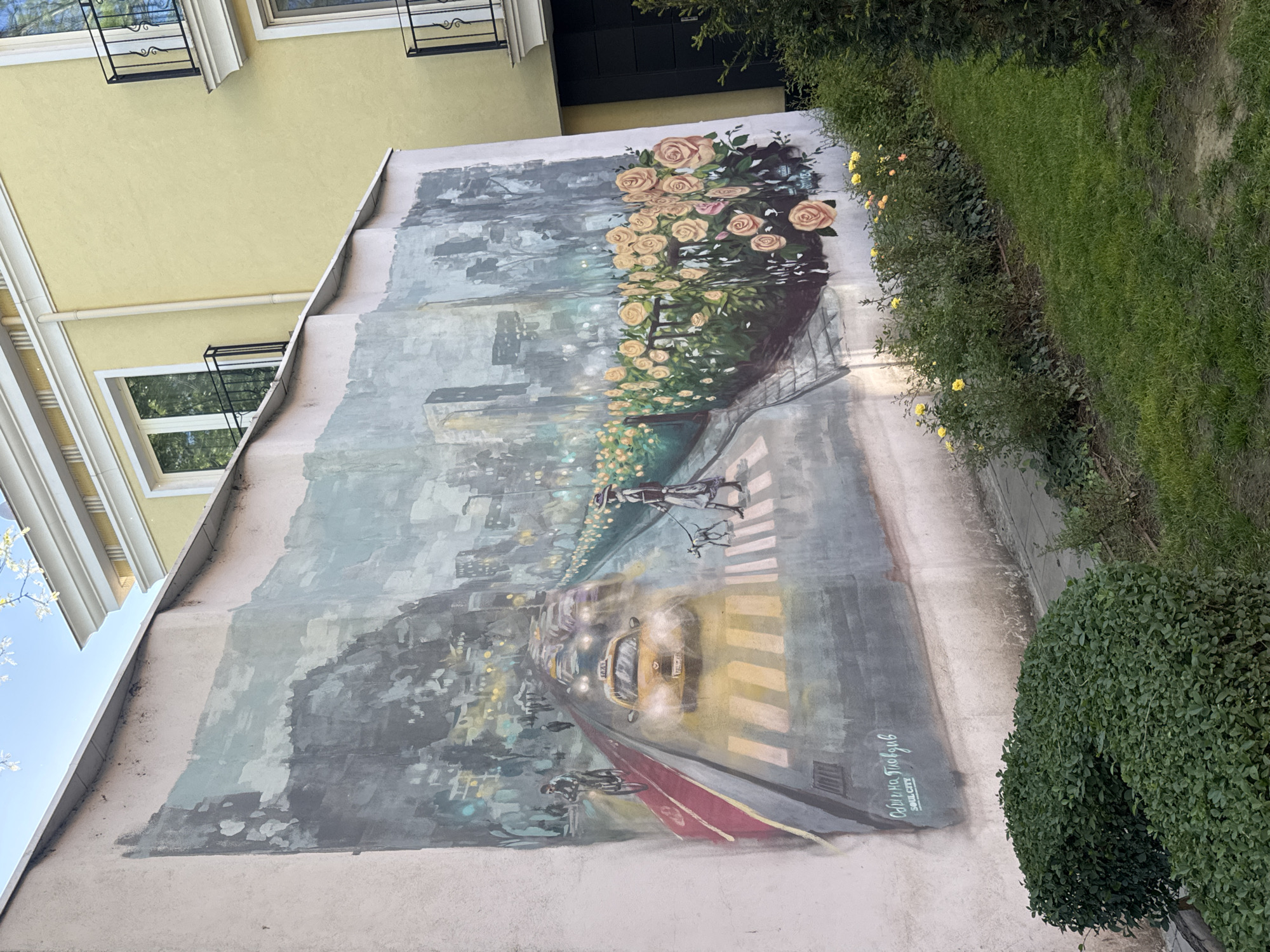The image shows a mural on the side of a building in Plovdiv, Bulgaria. The mural depicts a city scene, featuring a yellow taxi, pedestrian crossing, and skyscrapers in the background. Above the city scene are painted flowers. To the left of the mural is the facade of the building with windows and decorative elements. To the right of the mural is a hedge. The artist's signature is visible at the bottom right of the mural.
