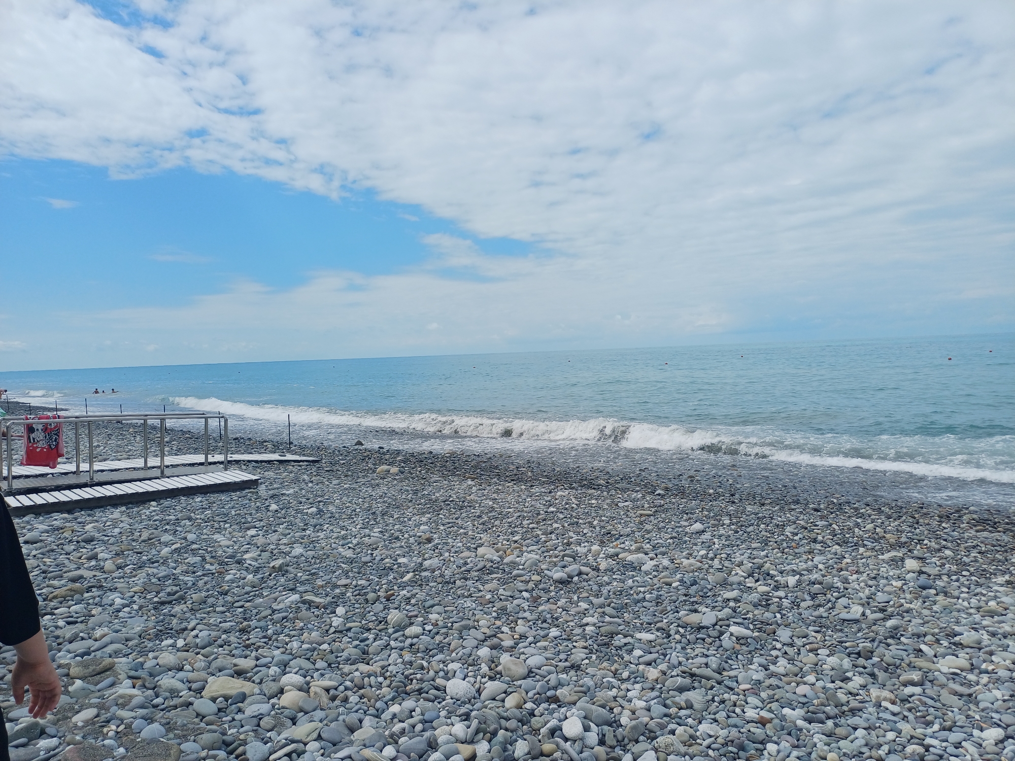 A bright, overcast day is depicted on a pebble beach in Sochi, Russia. The sky is a mixture of blue patches and dense white clouds. The sea is a calm, light blue, with small waves breaking gently on the shore. The beach is covered in smooth, grey and white pebbles. On the left side of the frame, a person's arm and hand are visible, suggesting they are walking towards the water. A metal railing and wooden planks form a walkway leading down to the beach. A red bin is positioned next to the walkway. In the distance, a few people are visible in the water, swimming and enjoying the sea.