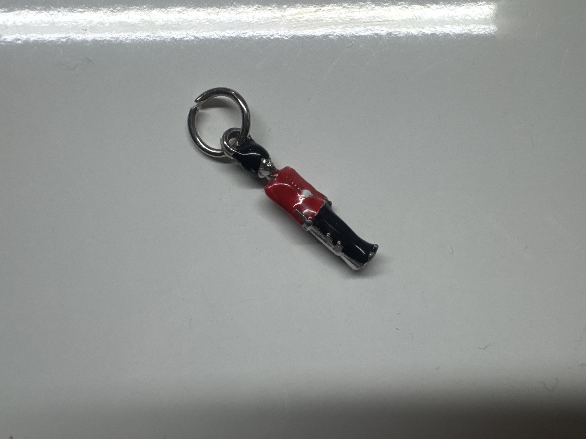 The image displays a small, stylized enamel charm, approximately 3-4 cm in length, positioned diagonally on a light grey, moderately reflective surface within the city of Unknown, Unknown. The charm depicts a figure with a red upper section, exhibiting visible white paint wear and metallic silver accents, and a black lower section with similar silver detailing. It is suspended from a small, silver-colored metal jump ring, which connects to a slightly deformed larger silver-colored metal split ring. The surface beneath the object contains scattered dust particles, fine scratches, and subtle smudges. A strong vertical reflection of light is present on the right side of the surface, indicating an external light source.