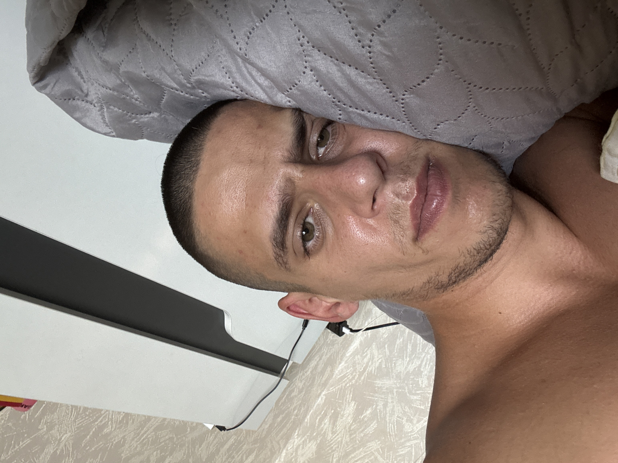 A close-up, upside-down, medium shot captures a fair-skinned man lying on his side with his head resting on a quilted gray pillow. He has short, dark hair, stubble on his chin and upper lip, and noticeable stubble on his eyebrows. His eyes are open and appear to be looking towards the camera. He has full lips, which are slightly parted. The man's neck and upper chest are visible, and he is wearing a light-colored garment. Behind him, a white piece of furniture with a dark gray surface is visible, and to the right, a wall with a textured wallpaper pattern is present. The overall lighting suggests an indoor setting, possibly a bedroom. There is no information in the image to determine the location in Sofia, Bulgaria.