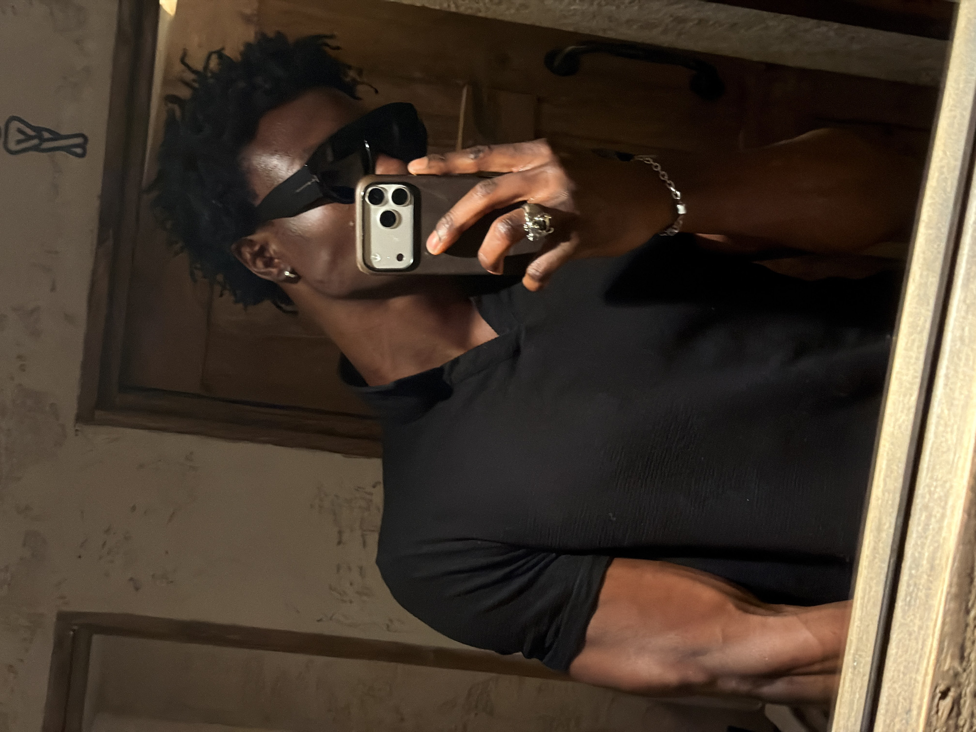 A dark-skinned man takes a selfie in a mirror. He wears dark sunglasses and a black shirt. The room's walls have a textured surface. A dark handle is visible on the wall behind him. A silver bracelet is on his left wrist, and a ring is on his left hand. The location is not discernible as being London based on the image provided.