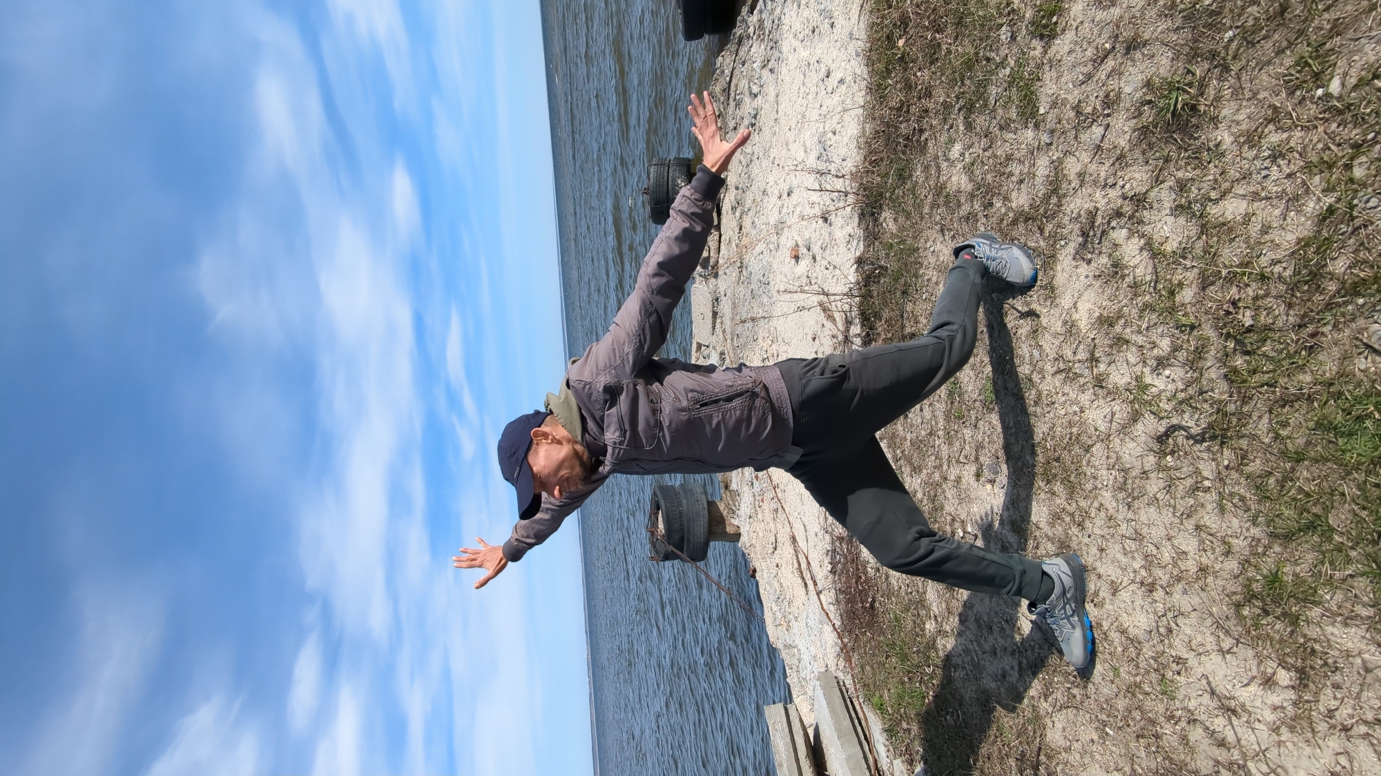 The image depicts a man playfully posing by a body of water under a bright, partly cloudy sky. The man, who has a beard and is wearing a dark baseball cap, a grey jacket over a lighter top, dark pants, and grey and blue athletic shoes, stands with his arms outstretched and his left leg bent upwards behind him, creating a dynamic, balancing pose. His expression suggests joy or relaxation.
The scene is an outdoor setting along a shoreline, with calm, blue-grey water visible on the left side of the frame. The ground on the right, where the man is standing, is composed of sandy dirt with sparse patches of dry, light green grass and some visible rocks. Several dark, worn-out tires are partially visible near the water's edge, possibly serving as part of a makeshift barrier or dock. The sky above is a clear blue with streaky white clouds, indicating good weather. The bright natural light suggests it is daytime. There is no visible text in the image.