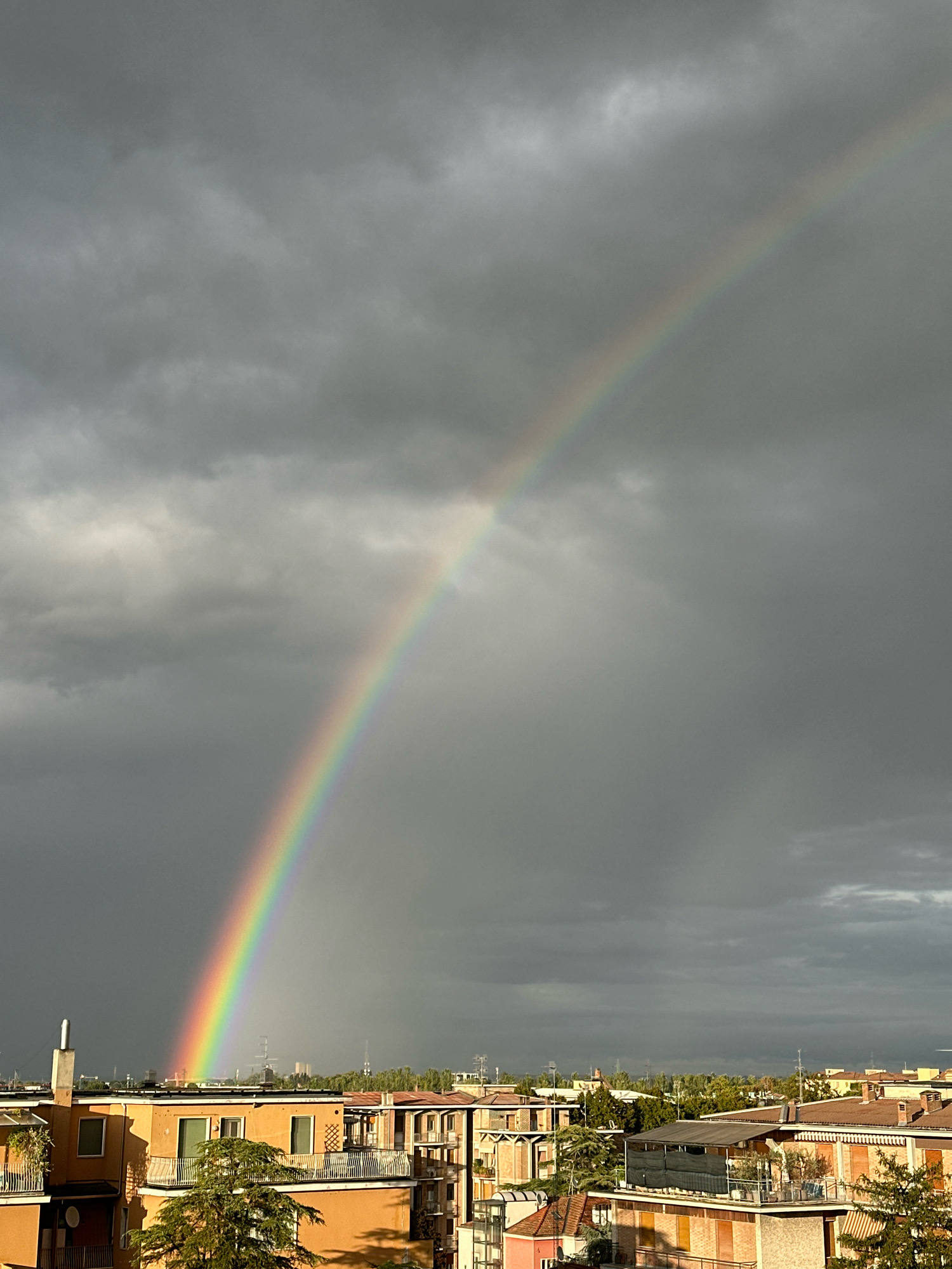 A rainbow arcs across a cloudy sky over a city. The sky is filled with dark, heavy clouds, suggesting recent or imminent rain. The rainbow, a bright spectrum of colors, stands out against the gray backdrop. Below the sky, buildings with orange and terracotta roofs are visible, indicating an urban environment. Trees and some foliage are interspersed among the buildings. In the distance, more buildings and possibly industrial structures can be seen. The lighting suggests it is daytime, likely late afternoon or early evening, given the dramatic cloud formations and the appearance of the rainbow. There are no people or discernible actions taking place in the image. This scene is consistent with the weather patterns observed in Parma, Italy.