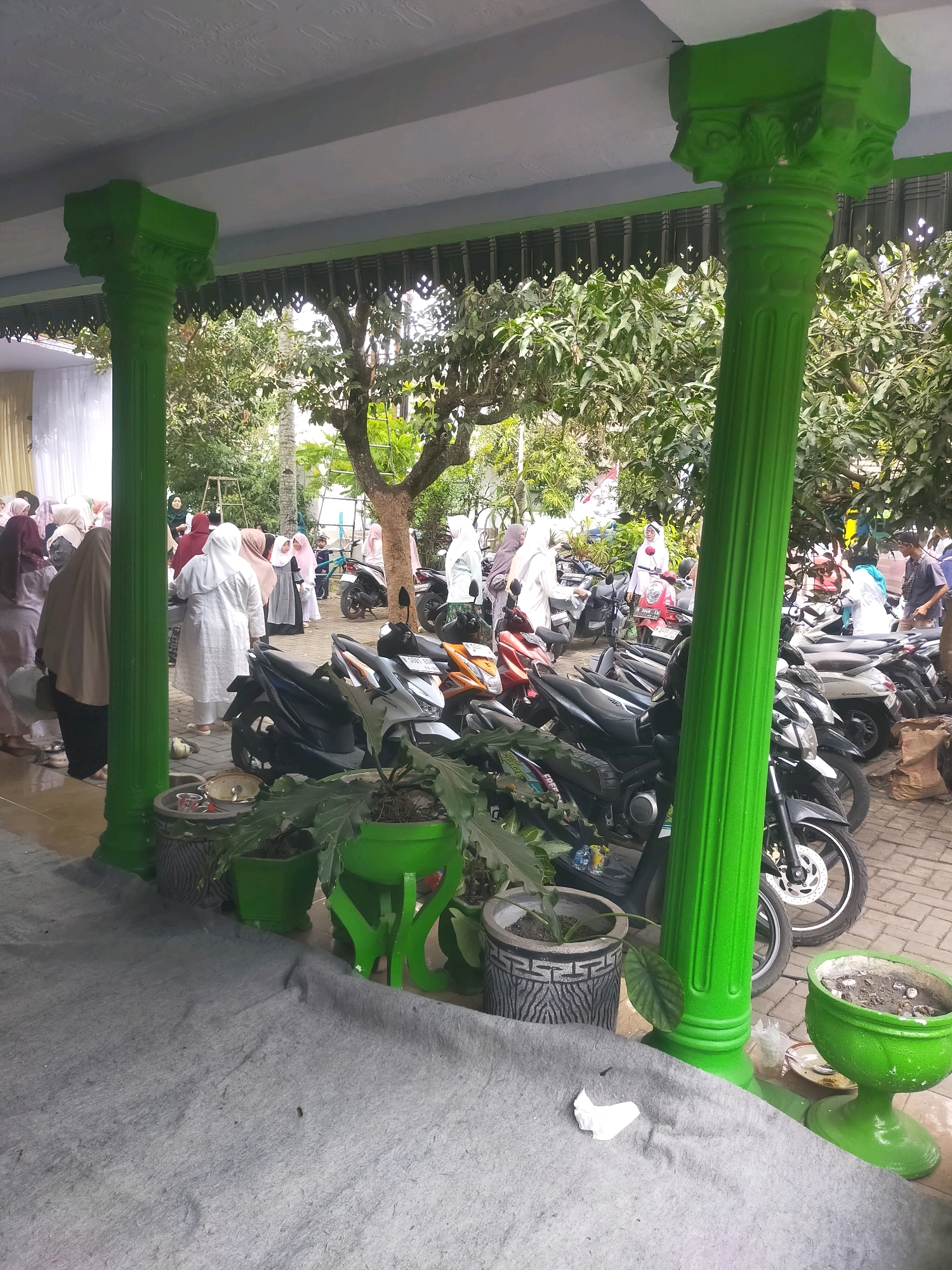 The image depicts an outdoor scene at what appears to be a communal gathering or event in Bululawang, Indonesia. In the foreground, two large, ornate green pillars stand prominently, supporting a covered walkway. A grey tarp covers a section of the ground in front of these pillars, upon which several potted plants are placed.
Behind the pillars, a variety of motorcycles are parked in neat rows, suggesting a transportation hub or a designated parking area for attendees. The motorcycles are a mix of colors and models, with some appearing to be scooters and others more standard motorcycle designs.
In the mid-ground and background, a group of people, predominantly women, are visible. Many are wearing hijabs and modest clothing, indicating a cultural or religious context. They are seen interacting and walking amongst themselves, with some near the parked motorcycles and others further back towards a white tent structure that suggests a venue for the event. Trees and foliage are abundant throughout the scene, indicating an outdoor environment with a significant amount of greenery. The ground surface appears to be paved with bricks in the area where the motorcycles are parked. The overall atmosphere suggests a daytime gathering, possibly a wedding, festival, or other community event.