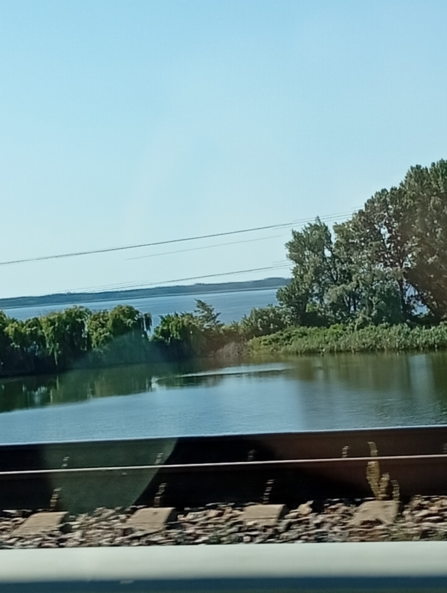 The image depicts a tranquil scene featuring a body of water bordered by lush green trees and vegetation. In the foreground, a railway track runs horizontally across the frame, indicating a transportation route. The sky above is clear and bright blue, suggesting a sunny day. The reflection of the trees and the sky on the water adds to the serene ambiance of the landscape. The overall impression is one of natural beauty and peacefulness, with man-made elements like the railway track subtly integrated into the environment.