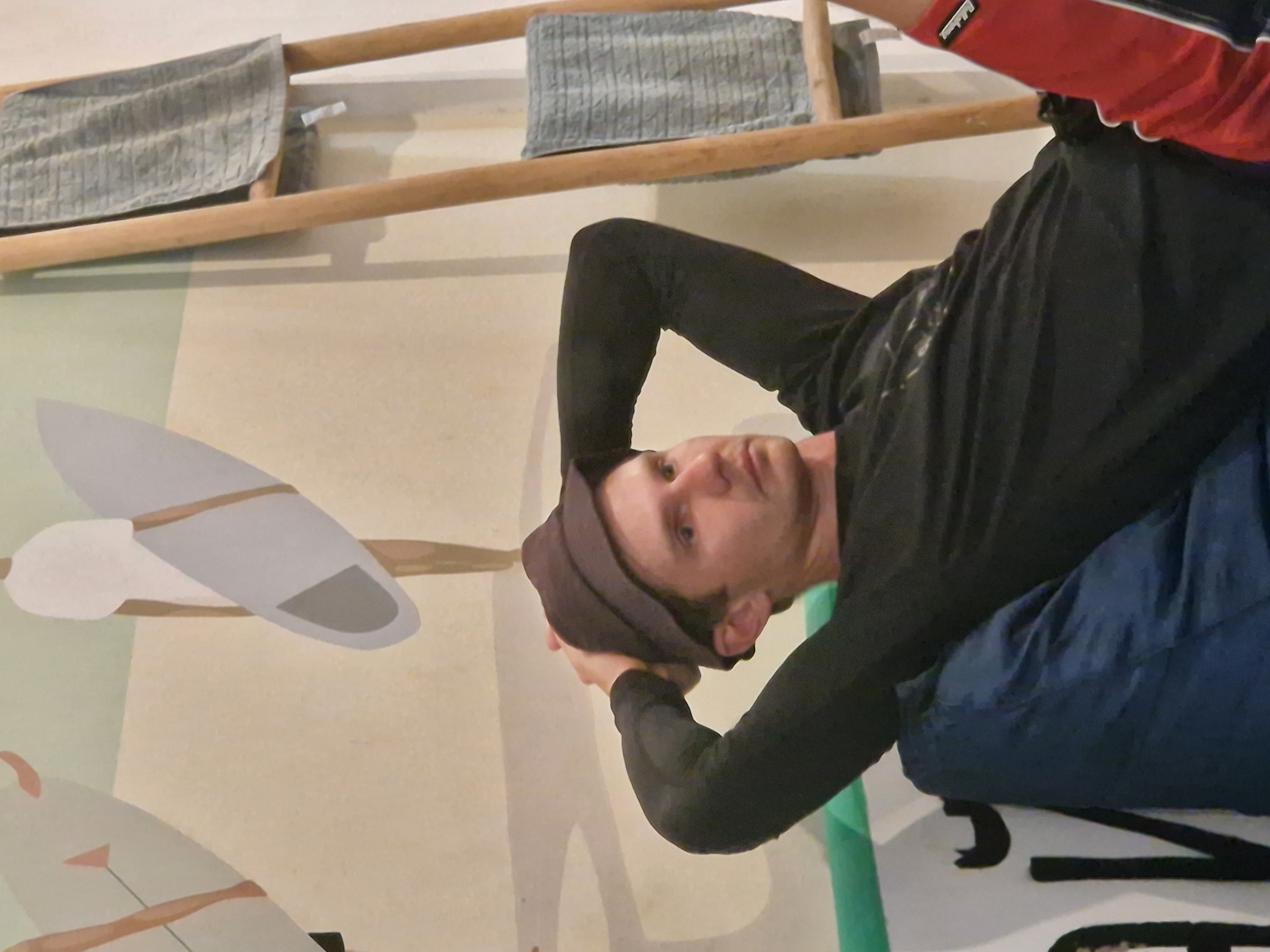 The image depicts a man in a black long-sleeved shirt, dark head covering, red and black sleeves, and blue pants. He appears to be lying down. The background features a wall with a painted mural, including depictions of airplanes and what appears to be a wooden ladder or frame with cloths draped over it. Without additional context or information in the image itself, it is not possible to confirm whether the photograph was taken in Kuta, Indonesia.