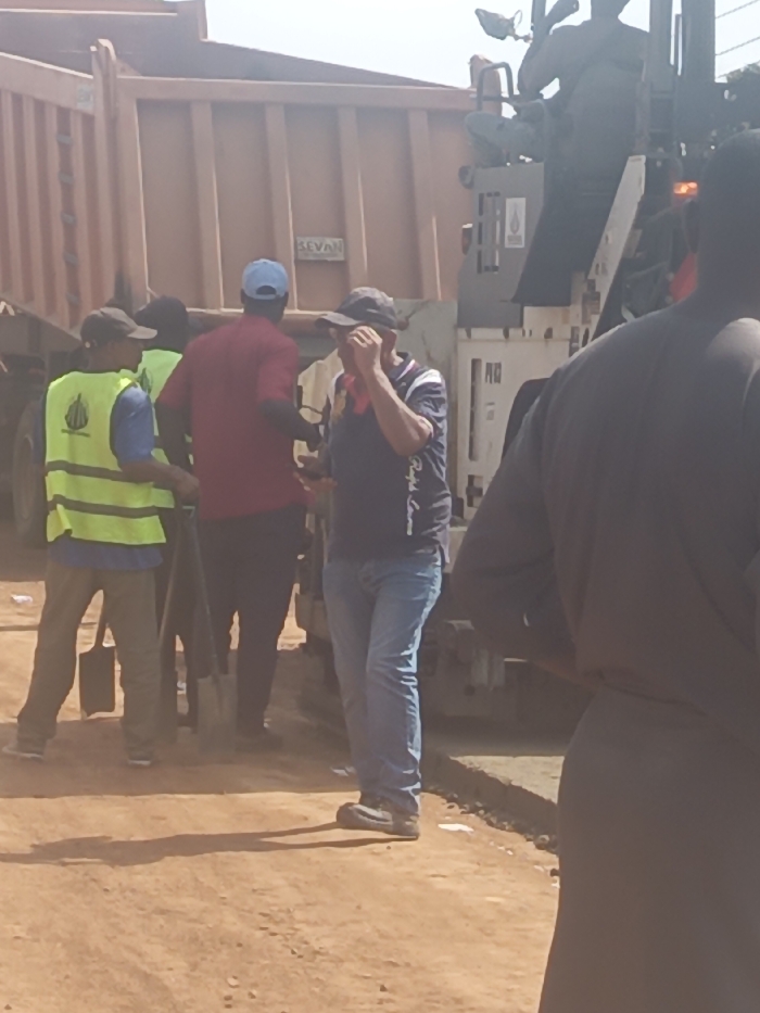 The image depicts an outdoor scene in Akwanga, Nigeria, characterized by construction or road work activity under bright daylight. In the background, a large orange dump truck with "SEVAN" visible on its side is parked. To the right of the truck, a heavy construction machine, possibly an asphalt paver, is present, with a person seated atop its upper deck. A "PMD" label is visible on the machine's side.
Several individuals are present on the unpaved, reddish-brown dirt ground. In the center-right, a man in a dark polo shirt with a "Ralph Lauren" logo, blue jeans, and a dark baseball cap raises his right hand to his face, seemingly wiping his eye or brow. To his left, slightly behind, a man wears a red short-sleeved shirt and a light blue baseball cap, facing away from the camera. Further left, a man is dressed in a blue shirt, a bright yellow-green safety vest with a visible logo, dark pants, and a dark baseball cap. He stands near two shovels leaning against the ground. The back of another person, wearing dark long-sleeved attire, partially obscures the foreground on the right. Shadows are cast on the dusty ground from the individuals and objects.
