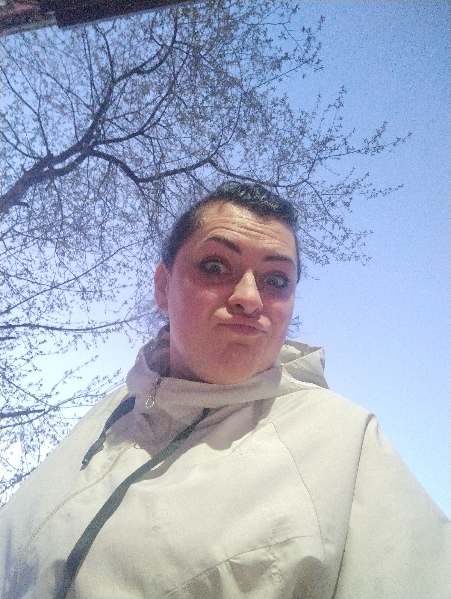 A woman takes a selfie outdoors. She has dark hair pulled back from her face, light skin, and wears a light-colored jacket with a hood pulled up around her neck. She is making a silly face with wide eyes and puckered lips. Behind her, a tree with bare branches spreads out against a pale blue sky. The exact location in Votkinsk, Russia, cannot be determined from the image alone.