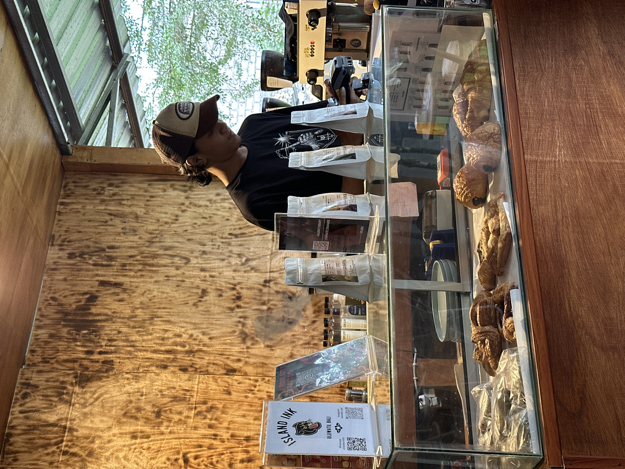 The image depicts a cafe counter in Kuta Selatan, Indonesia, likely a tattoo shop as suggested by the "Island Ink" sign. A young person wearing a black t-shirt and a brown and beige trucker hat is standing behind the counter, operating a card reader. They appear to be a barista or cashier. In front of them, on the glass-covered counter, are several bags of coffee beans or similar products, along with some pastries in plastic wrap and on display. The background features a wooden paneled wall and an espresso machine. Through a window or open space, trees and greenery are visible, indicating an outdoor or semi-outdoor setting. The overall atmosphere suggests a casual, local establishment.