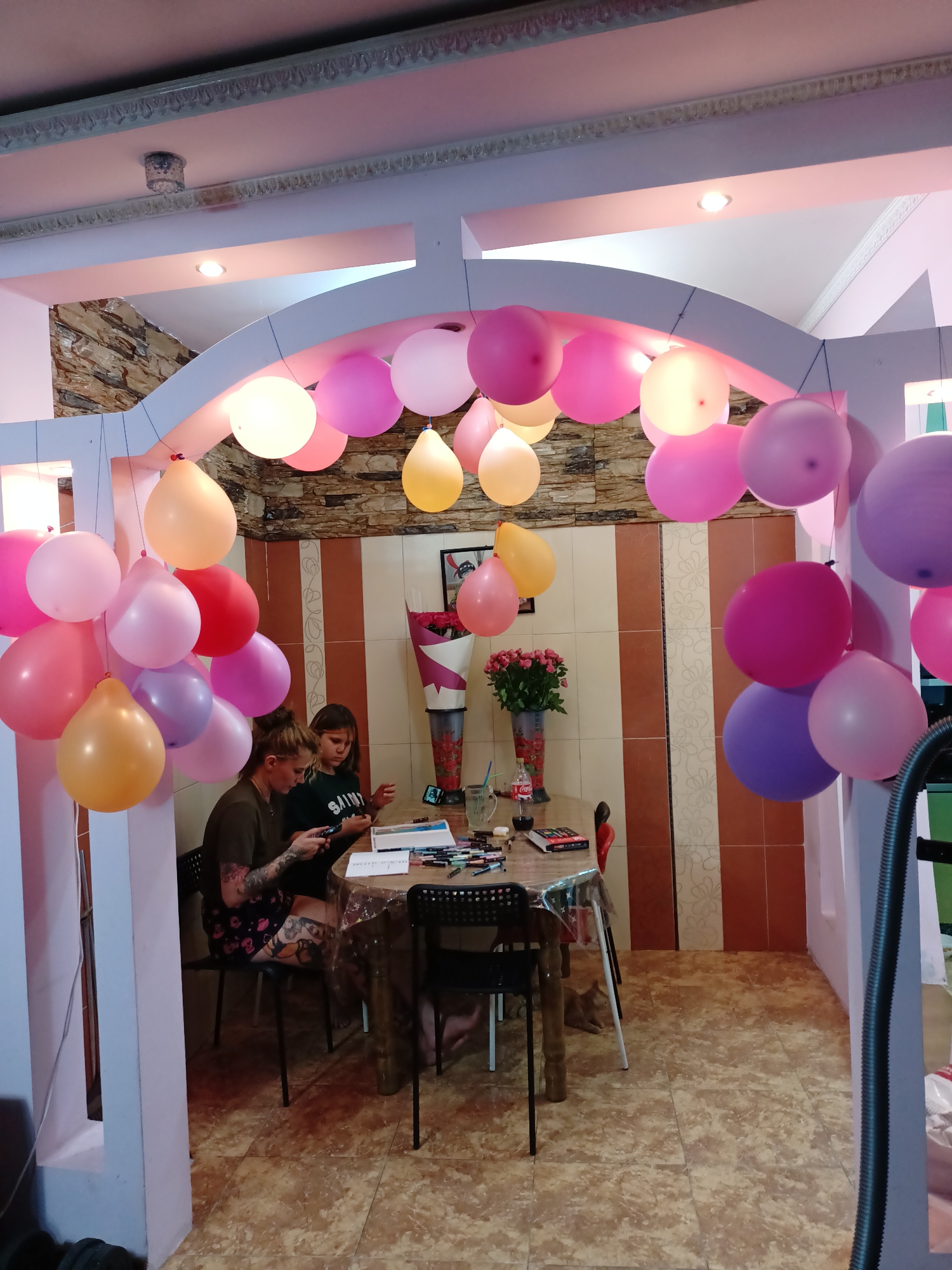 In a room decorated with numerous colorful balloons, two individuals are seated at a table. The individual on the left, a woman with tattoos on her arms and legs, is holding a smartphone. To her right, a young girl is looking down at the table, where art supplies are spread out. The table is covered with a plastic tablecloth adorned with a festive pattern. In the background, a vase filled with roses sits on the table, and a shelf holds a bottle of Coca-Cola and a glass. The room's walls feature striped tiling and a stone-like pattern. The overall ambiance suggests a celebration or a creative activity taking place. The image does not provide any specific landmarks or clues to definitively place the location in Almaty, Kazakhstan.