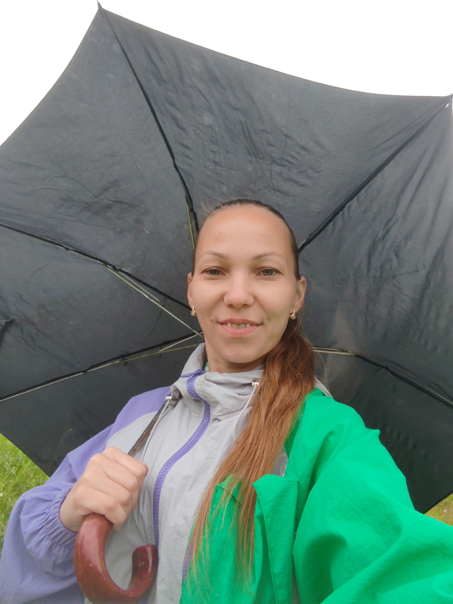 A woman is holding a black umbrella and smiling at the camera. She is wearing a light gray and purple jacket with a bright green hood. The umbrella is open and obscures the sky, suggesting it might be raining. The background shows a hint of green grass and possibly a cloudy sky. The image appears to be taken outdoors, in a non-urban natural setting, with no discernible elements pointing to the specific city of Чухлэм, Russia.