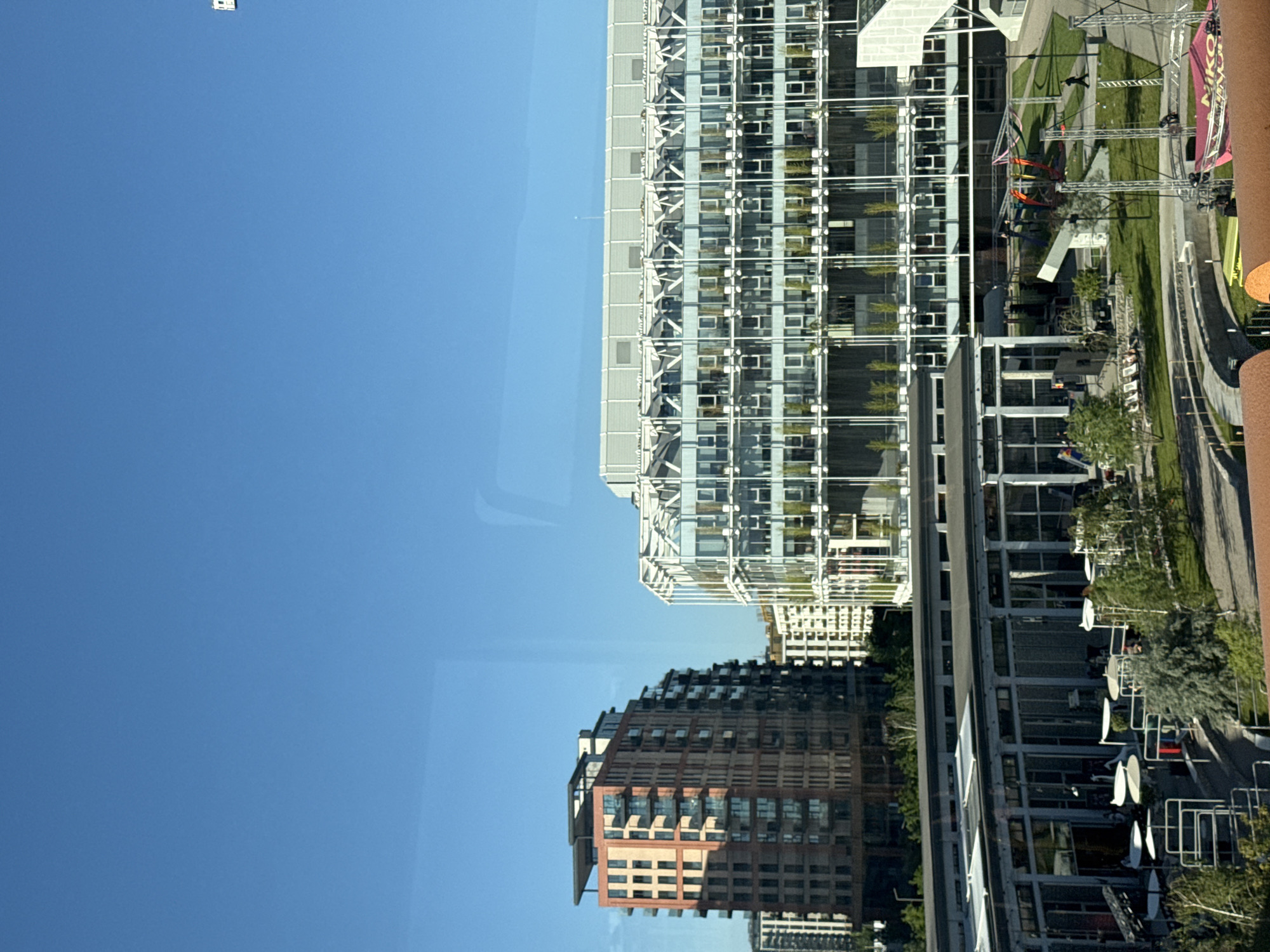The image captures an urban scene in Belgrade, Serbia. Two prominent buildings dominate the view: a tall, modern glass structure and a shorter, brick-clad building. The glass building appears to be a high-rise office or residential tower with numerous windows and balconies. The brick building displays a more traditional architectural style with a grid of windows.
Adjacent to the buildings is a landscaped area with trees, grass, and pedestrian walkways. There is also the visible presence of some objects, including scaffolding and signage. The clear blue sky fills the majority of the frame. The scene suggests a vibrant urban environment with a mix of modern and traditional architecture.
