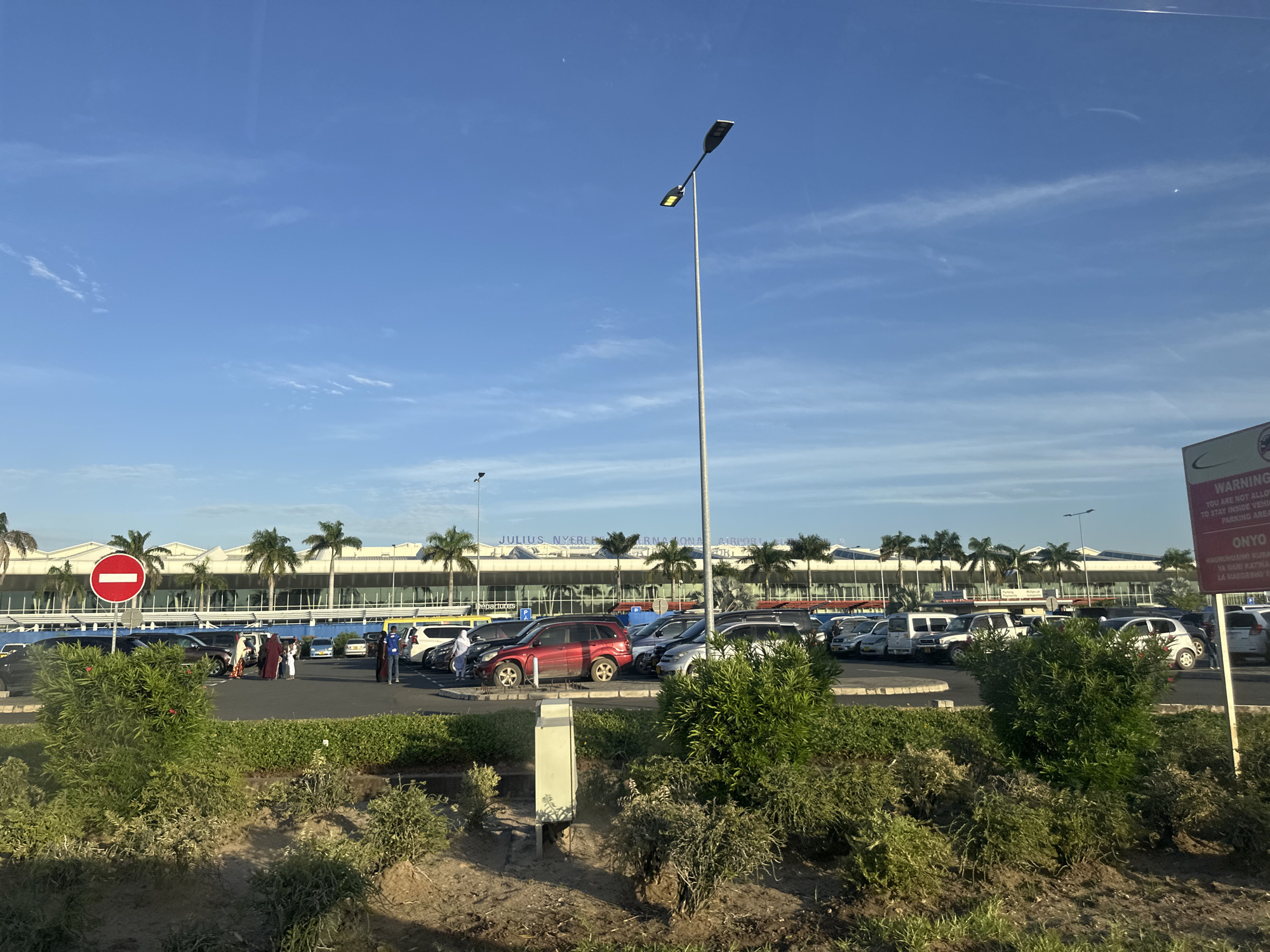 The image shows the front of Julius Nyerere International Airport in Dar es Salaam, Tanzania. A parking lot is situated in front of the airport building, populated with numerous parked cars. Several people are standing near the cars, some dressed in traditional Muslim attire. Palm trees line the front of the airport. Road signs are visible, including a no-entry sign and a warning sign about parking regulations. Bushes and grass cover the foreground. The sky is clear with a few wispy clouds.