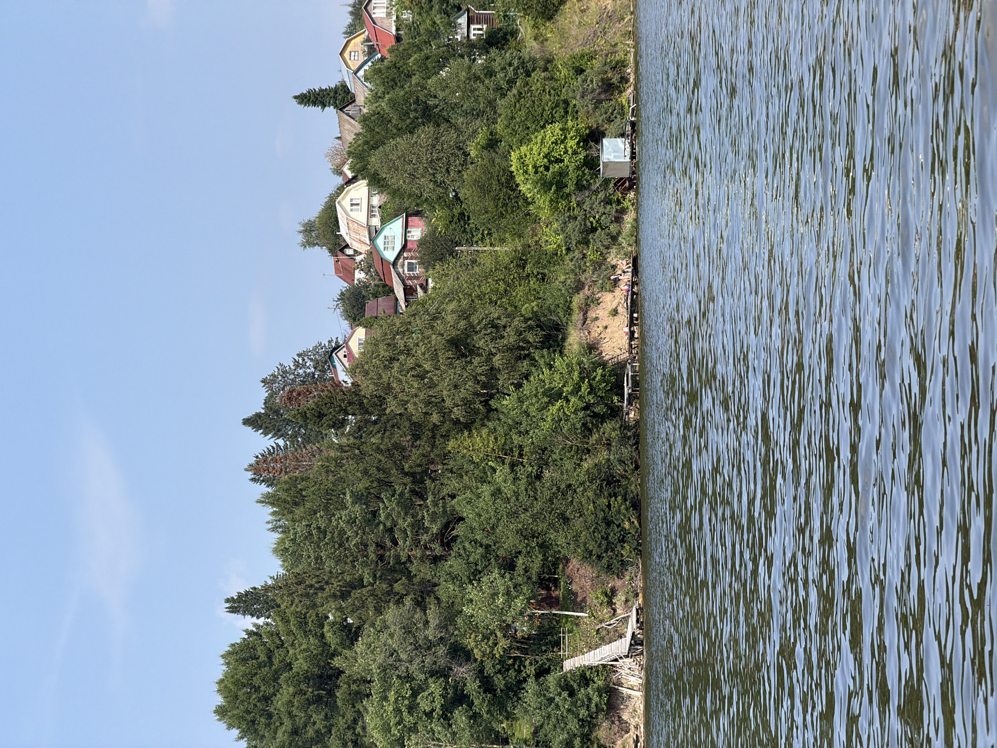 An overview shot shows a waterfront scene in Bykovka, Russia, on a clear day with a blue sky and some wispy clouds. The foreground is dominated by rippling water, likely a river or lake. In the middle ground, a green, tree-covered hillside rises steeply from the water's edge. A wooden pier extends into the water on the left, with a wooden ramp leading up the slope to a small wooded area. Further to the right, along the shore, are several wooden jetties, one of which supports a small, light-colored metal shed. A cluster of houses with colorful roofs is visible nestled on the crest of the hill, surrounded by trees. The houses are varied in design and color, with red, green, and yellow roofs being prominent. The trees are lush and green, with a mix of coniferous and deciduous species. The overall impression is of a peaceful, rural or semi-rural setting.