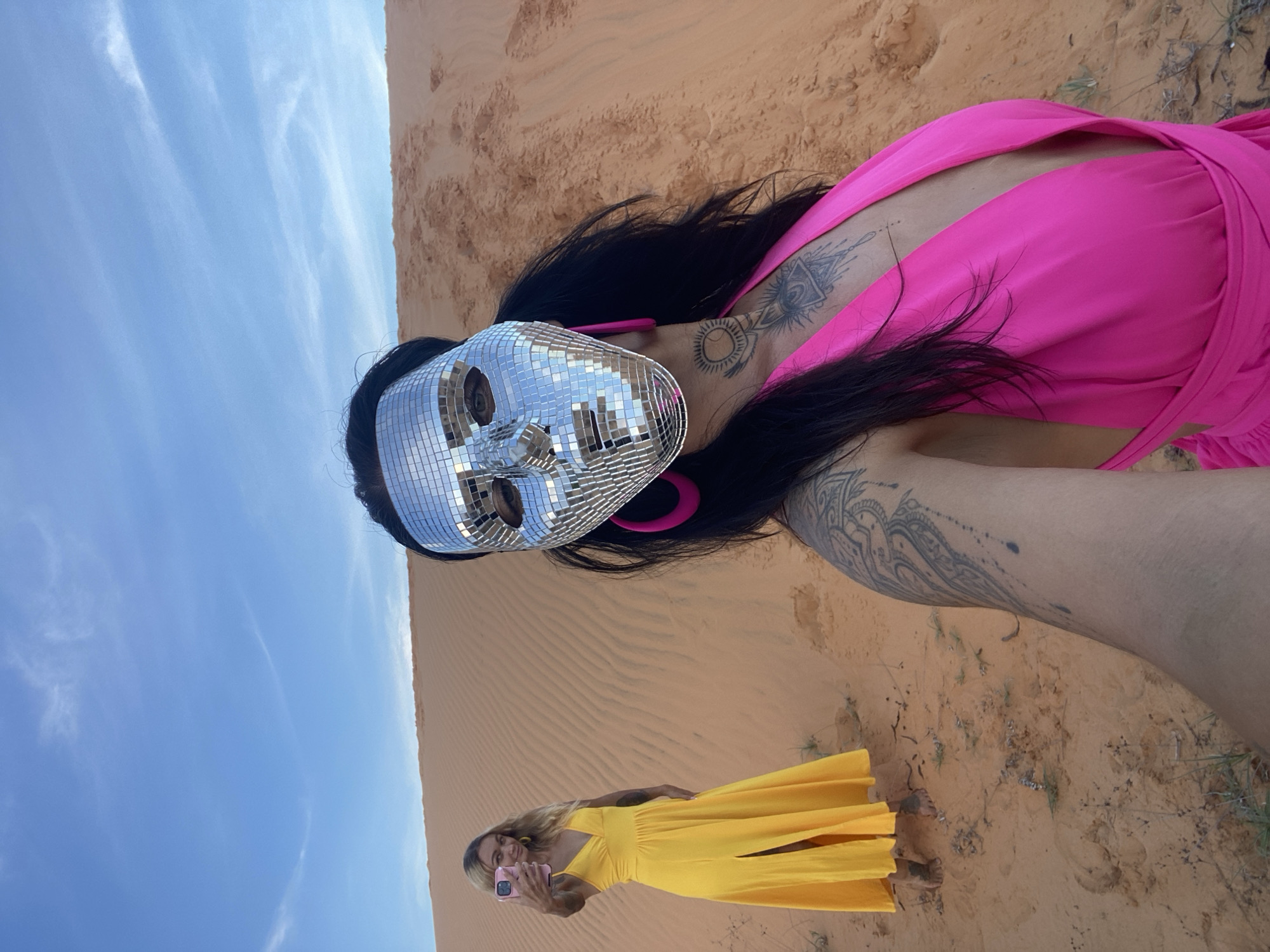 Two individuals are in a desert landscape under a blue sky with wispy clouds. The person in the foreground, positioned on the right, is wearing a bright pink low-cut outfit and a silver disco ball mask that covers their face. They have dark hair and visible tattoos on their chest and arm. Their right arm is extended, holding a phone, suggesting they are taking a selfie. Large pink hoop earrings are visible.
In the background, to the left, a second person is wearing a long, flowing yellow dress with a slit on the side. This person has blonde hair and is also holding a pink smartphone, seemingly taking a photo of themselves. They are barefoot. The environment consists of sand dunes with subtle textures and sparse, dry vegetation. The lighting suggests it is daytime. The location is identified as Mũi Né, Vietnam, a place known for its sand dunes.