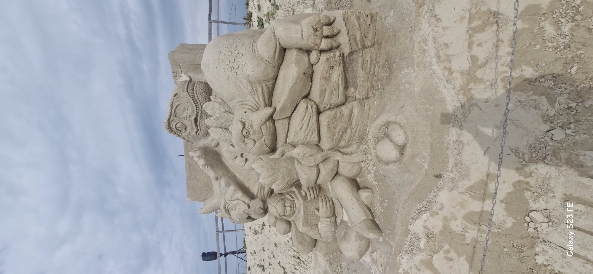 This image depicts a sand sculpture on a beach under a cloudy sky. The sculpture is complex, featuring several dinosaur figures and a human reading a book. In the foreground, a young person is seated, engrossed in reading a book titled "TREX." Beside them, a nest with two eggs is sculpted. Behind the person, large dinosaur figures are arranged, including what appears to be a Triceratops with horns and a Tyrannosaurus Rex with sharp teeth. Another smaller dinosaur, possibly a Stegosaurus with plates, is nestled among the larger ones. The overall scene evokes a prehistoric setting. In the background, a wooden fence runs along the beach, and a speaker on a tripod is visible to the left, suggesting an event or exhibition. The presence of chains in the foreground might indicate a boundary or safety measure. The location is stated to be the city of Jelgava, Latvia.
