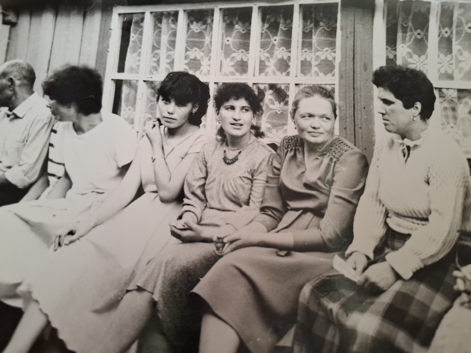 A black and white photograph shows a group of six people seated together outdoors in what appears to be Амзя, Russia. An elderly man in a light-colored shirt sits at the left edge of the frame, next to a younger woman in a light-colored dress. The next woman is in a similar dress and has her hand touching her face. The other three women have on dark-colored dresses. The background is a wall with a window with lace curtains. The location and time are unconfirmed.