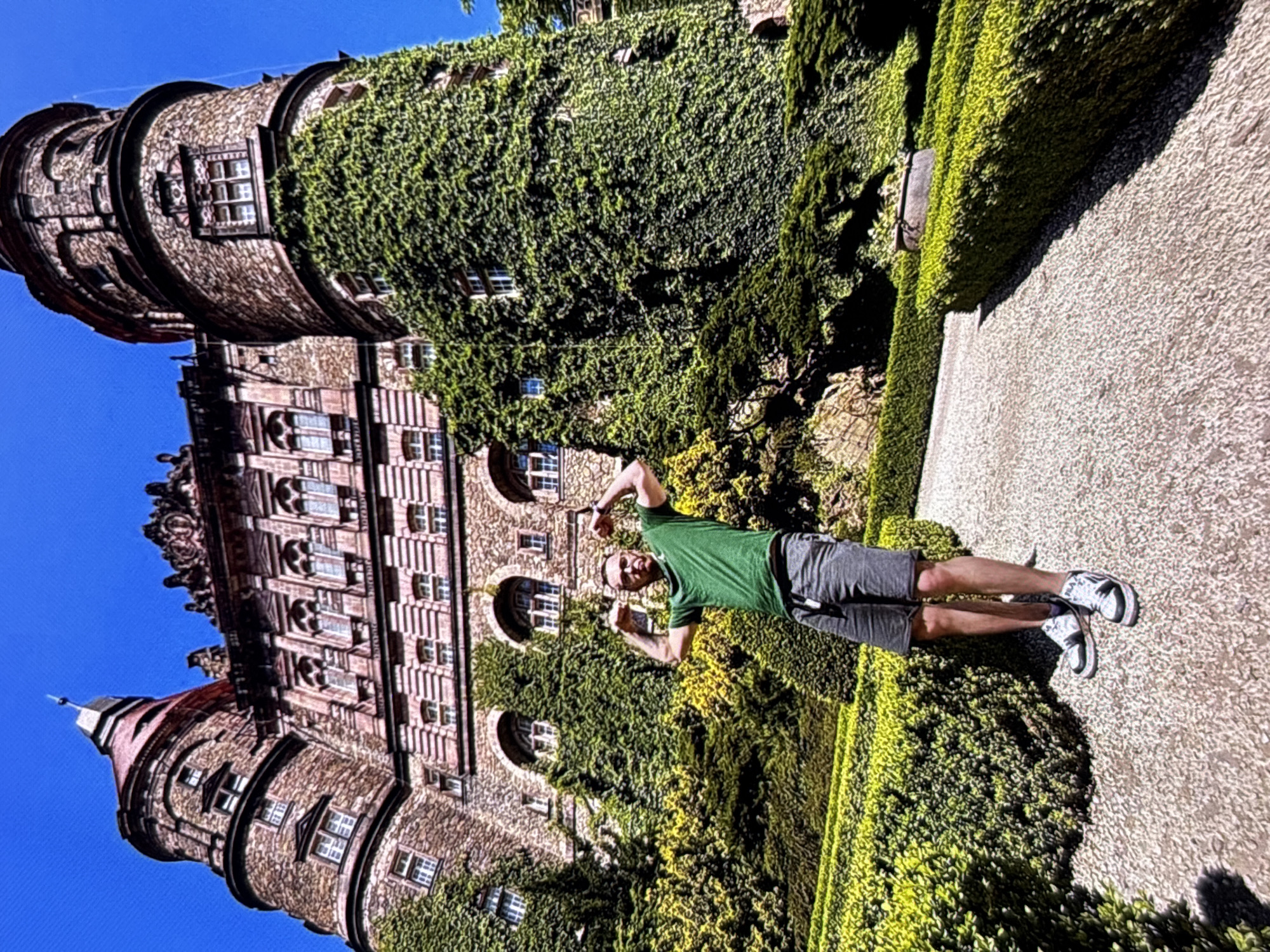 A man stands flexing his biceps in a garden in front of a large stone castle. The castle has multiple towers, some covered in ivy, and ornate architectural details. The garden features manicured hedges and a gravel pathway. The man is wearing a green t-shirt, grey shorts, and white and black sneakers. The sky is clear and blue. The location is the Książ Castle in Wałbrzych, Poland, not Radomsko.