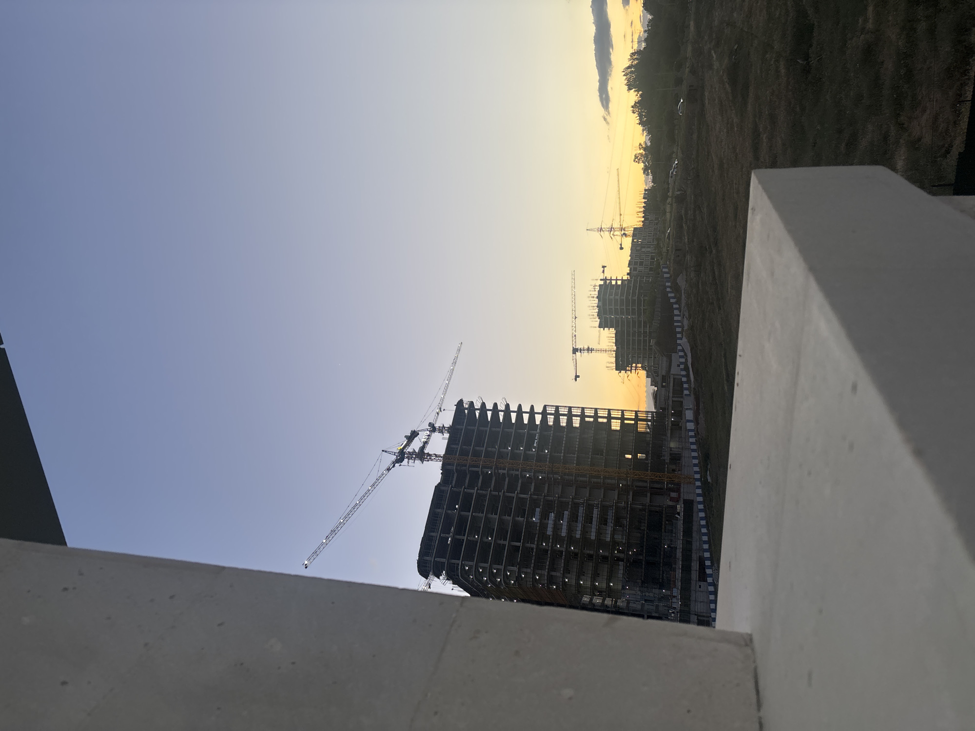The image, taken in Yerevan, Armenia, captures an expansive construction site at either sunrise or sunset. The sky displays a striking gradient, transitioning from a soft light blue on the left to a warm, bright yellow and orange near the horizon on the right, with a few faint clouds visible in the brighter portion.
In the midground, two large buildings are prominently under construction. The structure on the left, closer to the viewer, is a multi-story building with its concrete or steel skeleton exposed, showing multiple floors. A large tower crane extends from behind this building, its long arm reaching towards the sky. Further to the right, another building in a similar stage of development is visible, also surrounded by at least two additional cranes.
The right side of the background features a patch of dry, grassy terrain or a low hill, with a line of trees silhouetted against the bright horizon. Several vehicles are faintly discernible on this land. The foreground of the image is partially framed by concrete architectural elements, suggesting the viewpoint is from a building or balcony. No people or active construction work are visible, conveying a quiet, transitional moment of the day. There is no visible text in the image.