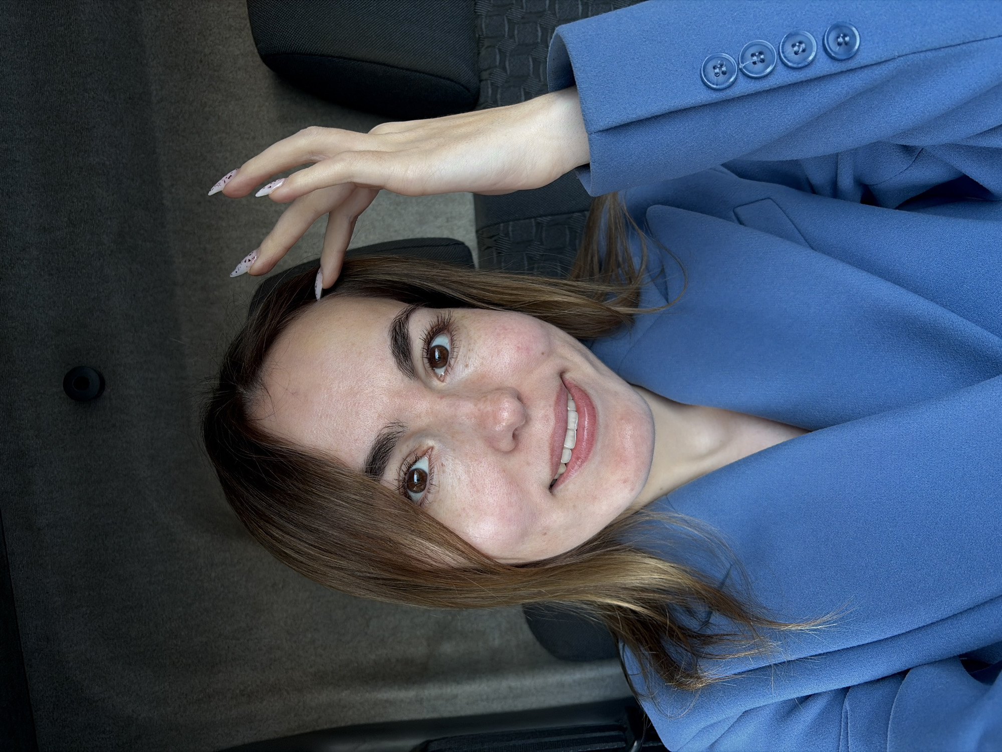 A female individual is positioned for a photograph inside a vehicle. She has light skin, brown eyes, and shoulder-length brown hair. Her mouth is open in a smile, revealing teeth. She is wearing a blue blazer, characterized by a visible lapel and a sleeve cuff adorned with four round blue buttons. Her right hand is raised, with the index finger touching the right side of her forehead near the hairline. Her fingernails are long, pointed, and light in color with dark, speckled patterns. The background shows the interior of a vehicle, including a dark grey upholstered ceiling (headliner) with a single black circular fixture visible in the upper left, and a dark grey textured fabric seat back and headrest to the right of the subject. The image does not contain visual information to independently confirm the provided location of Izhevsk, Russia.