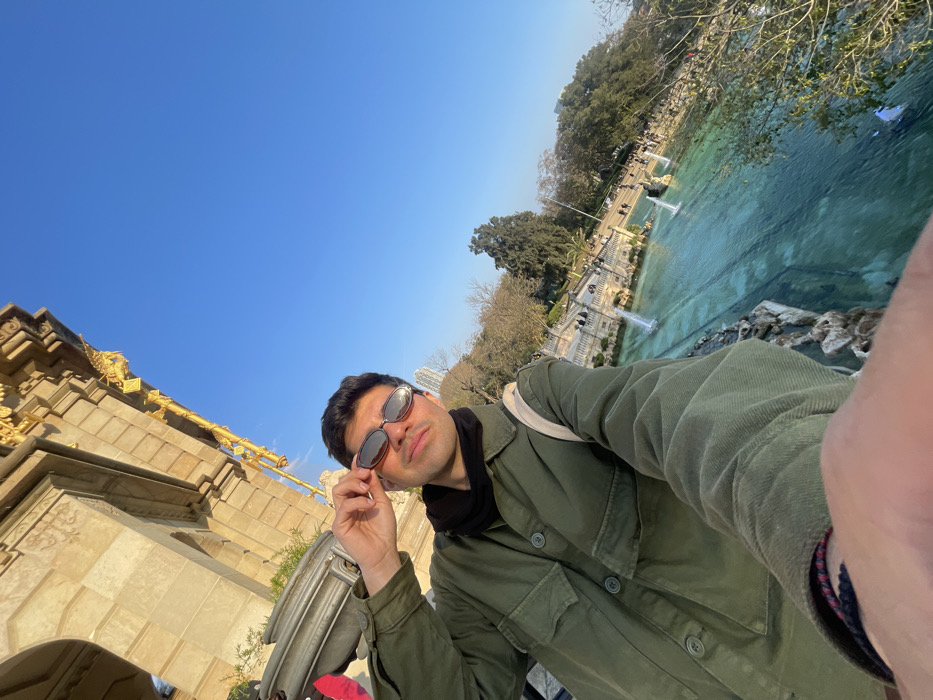 A young adult male, facing forward and slightly to the right, takes a selfie at the Cascada Monumental (Waterfall) in Parc de la Ciutadella, Barcelona, Spain. He wears an olive-green corduroy jacket, a black neck covering, and oval sunglasses, which he is adjusting with his left hand. A multicolored bracelet is visible on his left wrist. The background features the ornate golden and light stone architecture of the Cascada Monumental on the left, and a large turquoise water basin with active fountains on the right. Beyond the water, a landscaped park with trees, pathways, and pedestrians is visible under a clear blue sky. The scene is brightly illuminated by natural sunlight, suggesting a late afternoon or early morning setting.