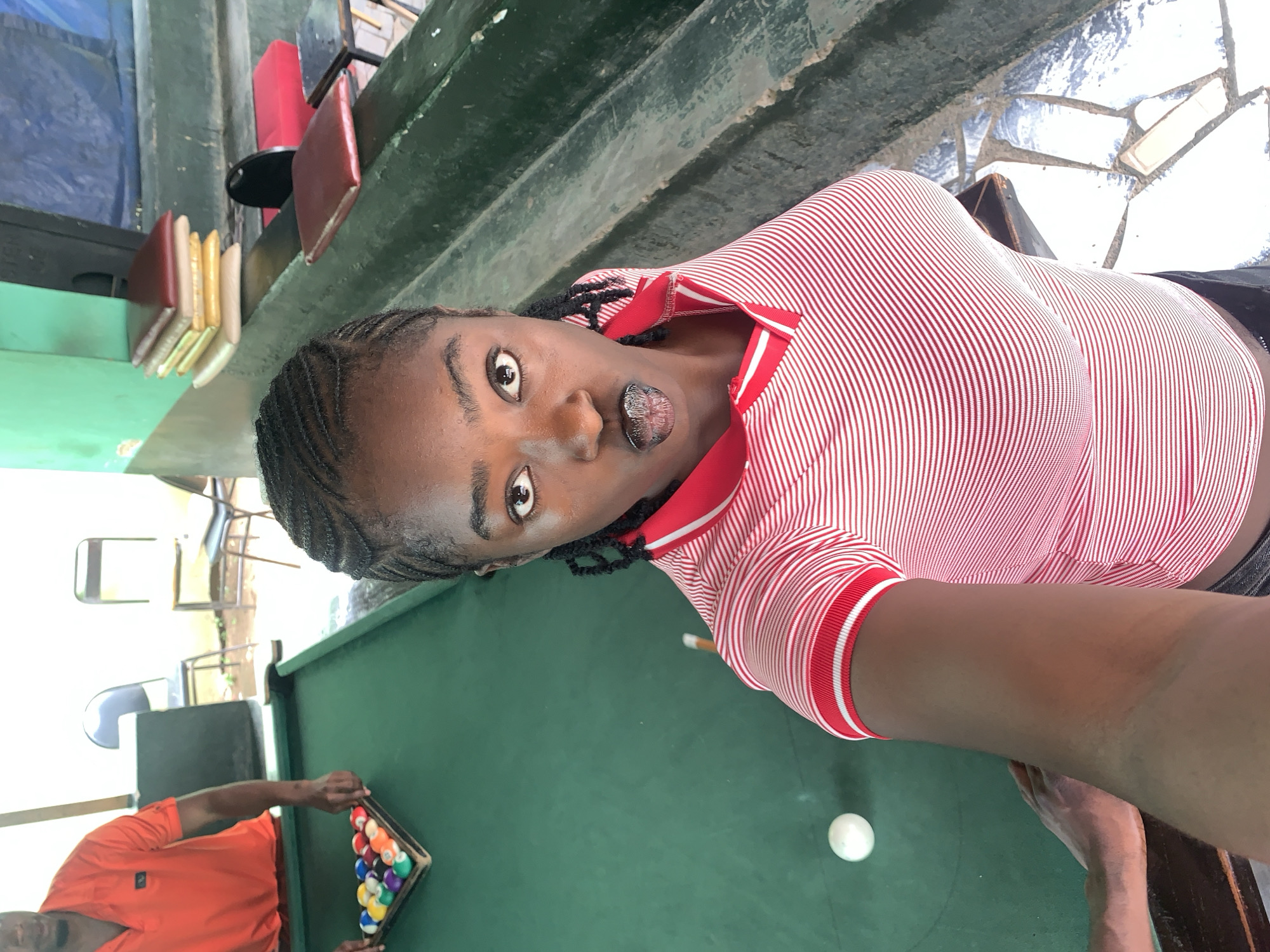 This portrait-oriented image depicts an indoor scene at a recreation area, likely within New Stadium, Nigeria. The foreground is dominated by a dark-skinned woman who appears to be taking a selfie or video. She looks directly at the camera with wide, expressive eyes and slightly parted lips, wearing a red and white striped polo shirt with a red collar and striped cuffs. Her hair is styled in dark braids.
The main activity centers around a green pool table that occupies a significant portion of the lower frame. To the lower left, a dark-skinned man in an orange t-shirt is visible, engaged in setting up a game. He is holding a wooden triangle rack containing a full set of colorful pool balls, with a white cue ball resting on the table.
The background shows various elements of the indoor setting: green walls or structures, red chairs, and a floor with irregular white and grey tiles. On a shelf or ledge in the upper background, several items resembling books with red and gold covers are stacked. Other chairs are visible further back. The overall atmosphere suggests a casual social space, possibly a pool hall or lounge, during daytime hours, as indicated by the ambient lighting. There is no visible text within the image.