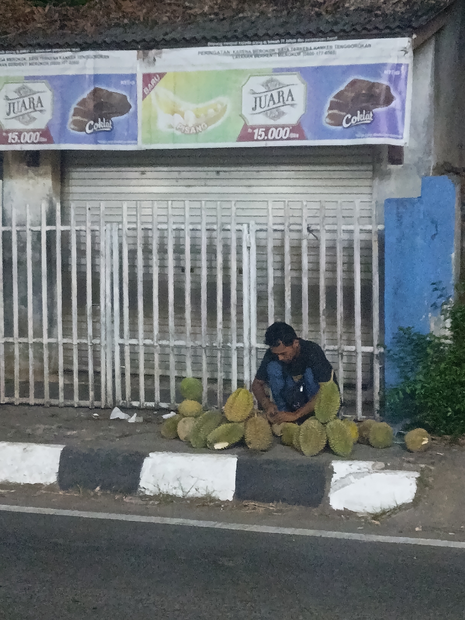 The image shows a man sitting on the ground in front of a metal gate, surrounded by durians. He is wearing a dark t-shirt and blue jeans. The durians are piled around him, and some are visible in the foreground. Behind the gate, there is a building with advertisements on the upper part. One advertisement features the word "JUARA" and a price of "15.000". Another advertisement shows a bunch of bananas with the word "PISANO". The scene appears to be outdoors, possibly a street or a marketplace, in Jonggol, Indonesia. The overall atmosphere is dimly lit, suggesting it might be dusk or evening.