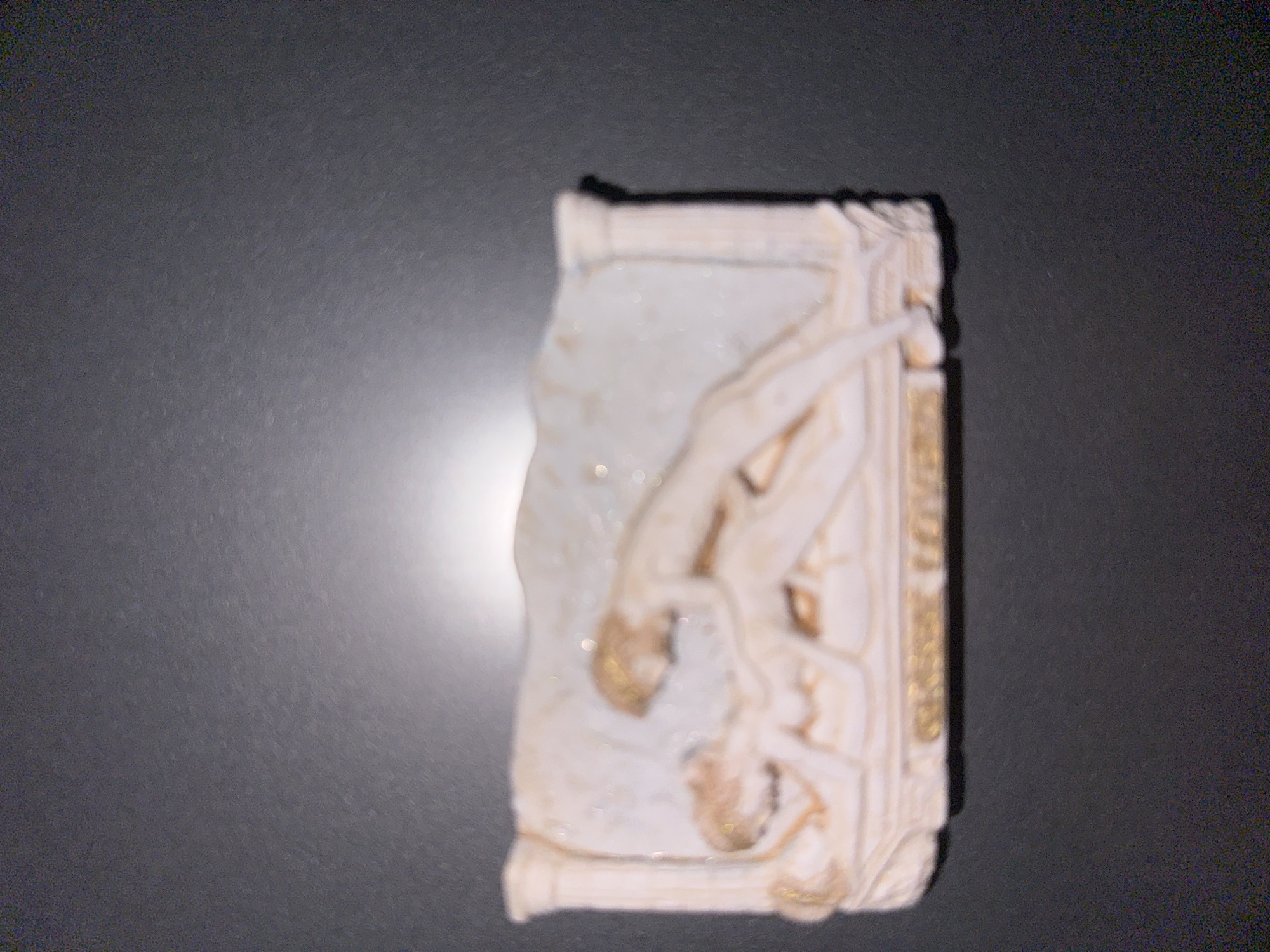 The image shows a relief sculpture of two nude figures in an embrace, suggestive of sexual activity. The sculpture is predominantly white with gold accents, and it is mounted on a dark, textured surface. Below the figures, there is an inscription in gold lettering that appears to read "SECRET LOVERS". The lighting in the image is uneven, with a bright light source casting a glow on the sculpture from the upper right. The sculpture is positioned horizontally in the center of the frame. The background is dark and out of focus, suggesting it is a wall or a similar surface. The image is blurry overall, but the central figures and the inscription are discernable. No information about the location in Ruse, Bulgaria is provided or inferable from the image itself.
