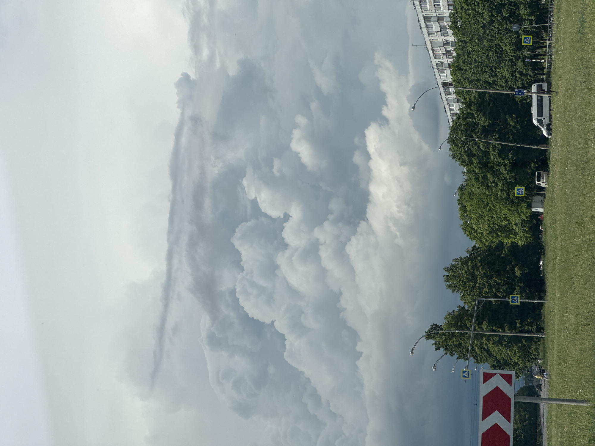 The image shows a landscape with an expansive sky filled with mammatus clouds. On the right side of the image, there is a strip of green grass, followed by a row of trees and buildings. Streetlights and road signs are also visible. A white van is parked near the trees. The scene appears to be in an urban area, possibly near a park or residential zone.