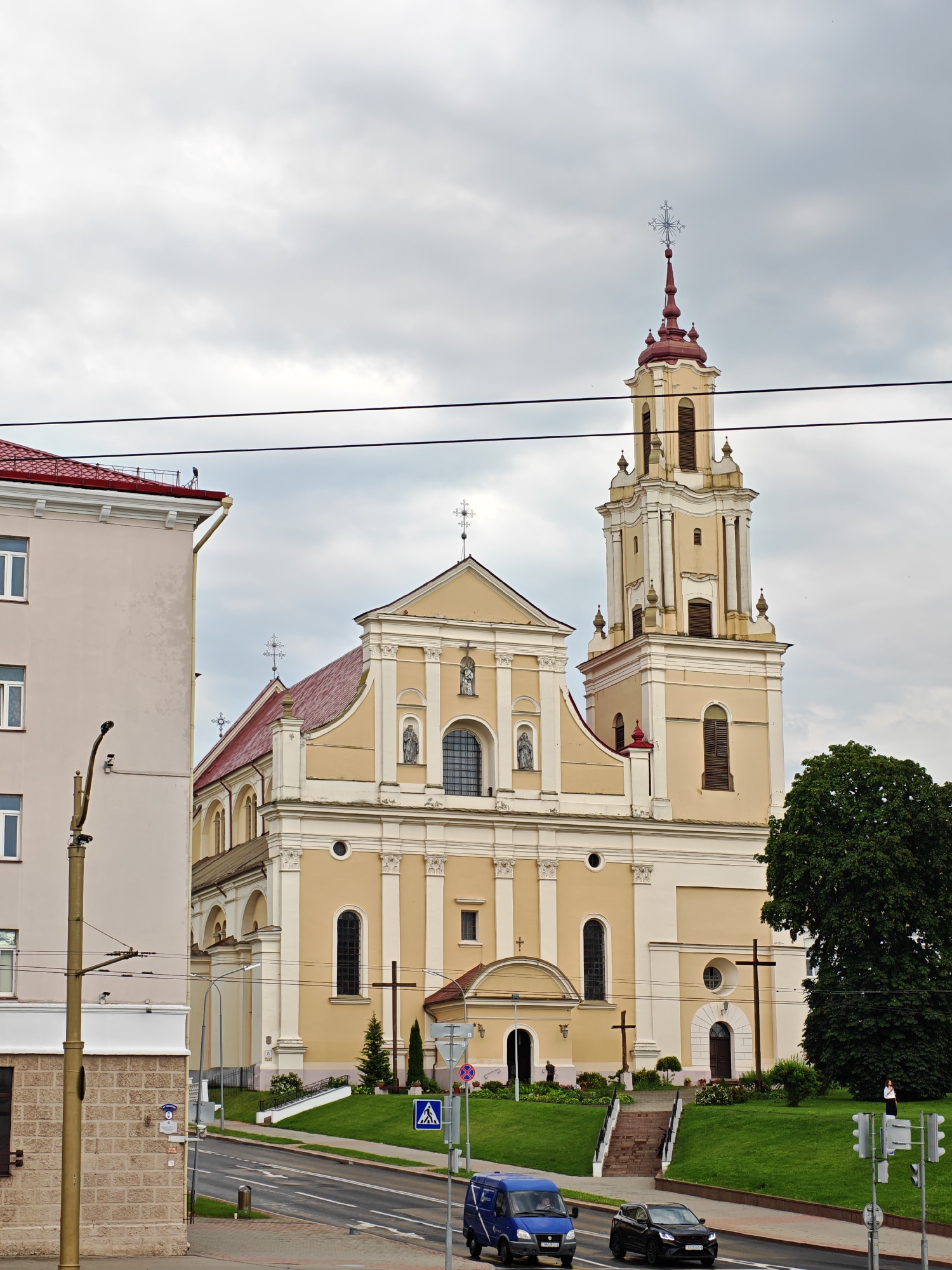 The image shows the Cathedral of St. Francis Xavier in Hrodna, Belarus, under a cloudy sky. The yellow-colored cathedral features a tall, multi-tiered bell tower with a red roof and a decorative cross at its apex. Multiple crosses are visible on the church facade and in the surrounding grounds. A light-colored building with a red roof is partially visible on the left, adjacent to the cathedral. A street runs in front of the cathedral, with a blue van and a black car driving on it. There are traffic lights, street signs, and a lamppost visible along the street. The foreground shows a grassy area with a few trees and some steps leading up to the cathedral entrance. A few people are visible in the image, including two near the entrance of the cathedral and one standing on the grassy area to the right. Electrical wires are prominent in the upper portion of the image.
