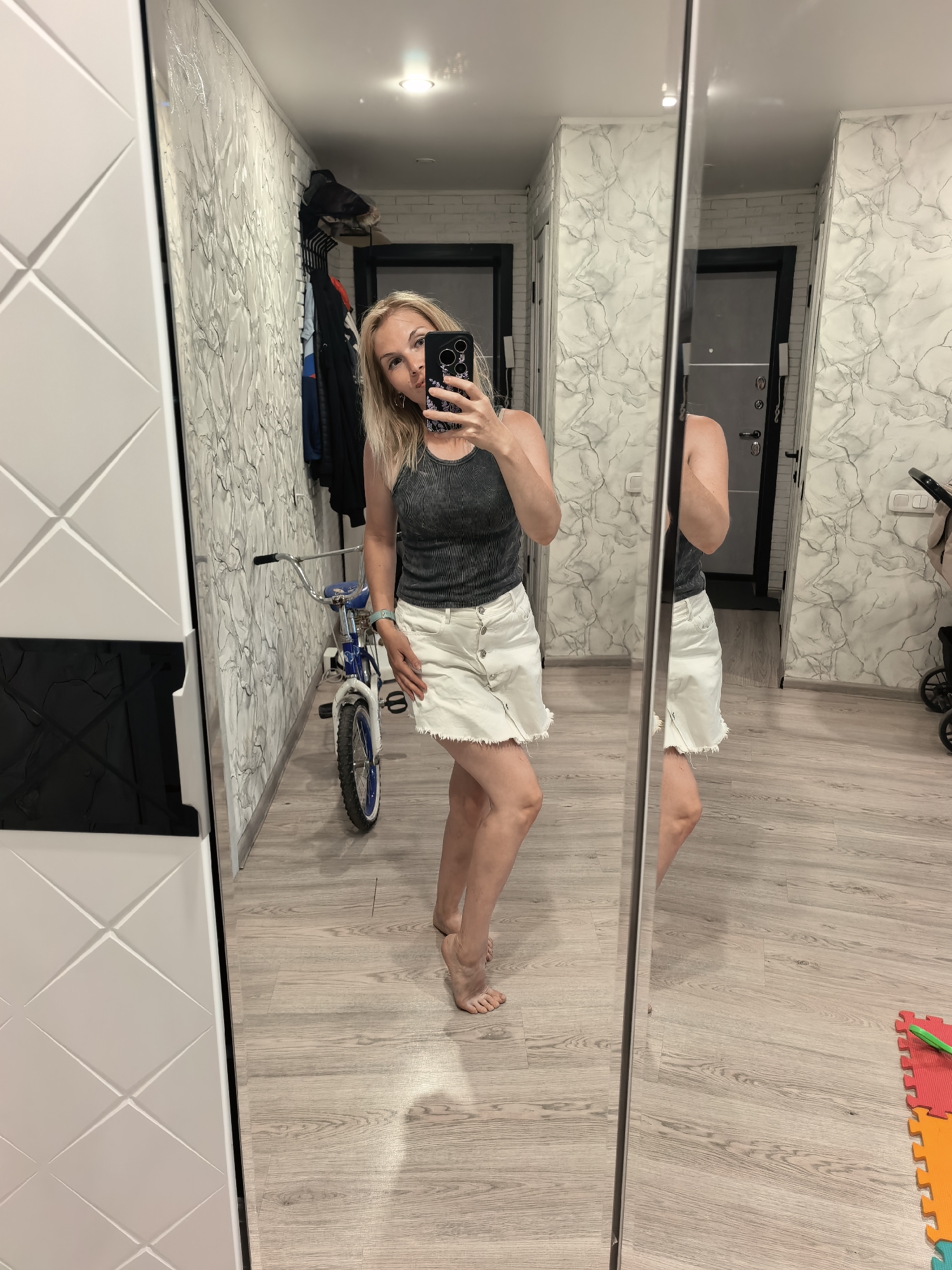 A woman is taking a mirror selfie in what appears to be an apartment. She is wearing a dark tank top and a short, white button-down skirt. The room has light-colored walls with a marble pattern, light wood-look flooring, and white built-in cabinets. A child's bicycle is visible near the wall, along with clothing hanging in the background. A stroller and children's play mats can also be seen, suggesting a family residence. The location is Sterlitamak, Russia.