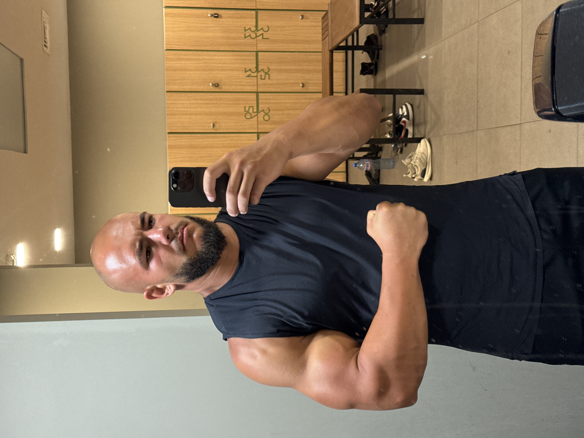 A bald, bearded man in a sleeveless dark t-shirt and black shorts is taking a mirror selfie in a gym locker room. He is flexing his left bicep. In the background, there are wooden lockers and benches. A water bottle and various athletic shoes are scattered on the floor. The floor is tiled. The lighting appears to be overhead fluorescent lights. The overall atmosphere is that of a typical gym environment.