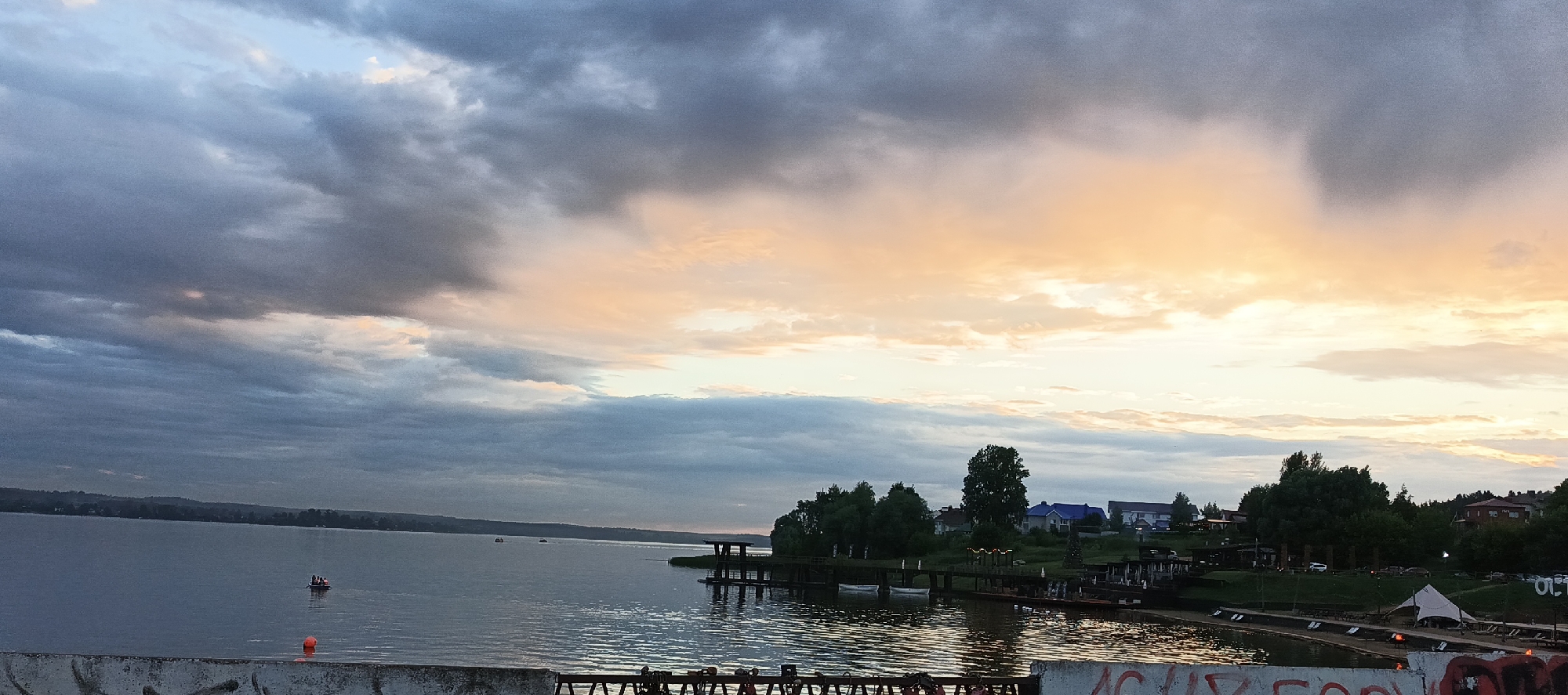The image depicts a panoramic view of a body of water, likely a lake or river, in Votkinsk, Russia. The sky is filled with dramatic clouds, transitioning from dark grey on the left to soft hues of peach and yellow on the right, suggesting either sunrise or sunset. The water's surface is calm, reflecting the sky's colors.
Along the far bank of the water, a treeline forms a natural border, interspersed with houses and buildings. A small dock structure extends into the water from the right bank. There are some boats visible on the water. In the foreground, there is a concrete structure with graffiti on it, possibly a bridge or embankment. The overall scene conveys a sense of tranquility and natural beauty.