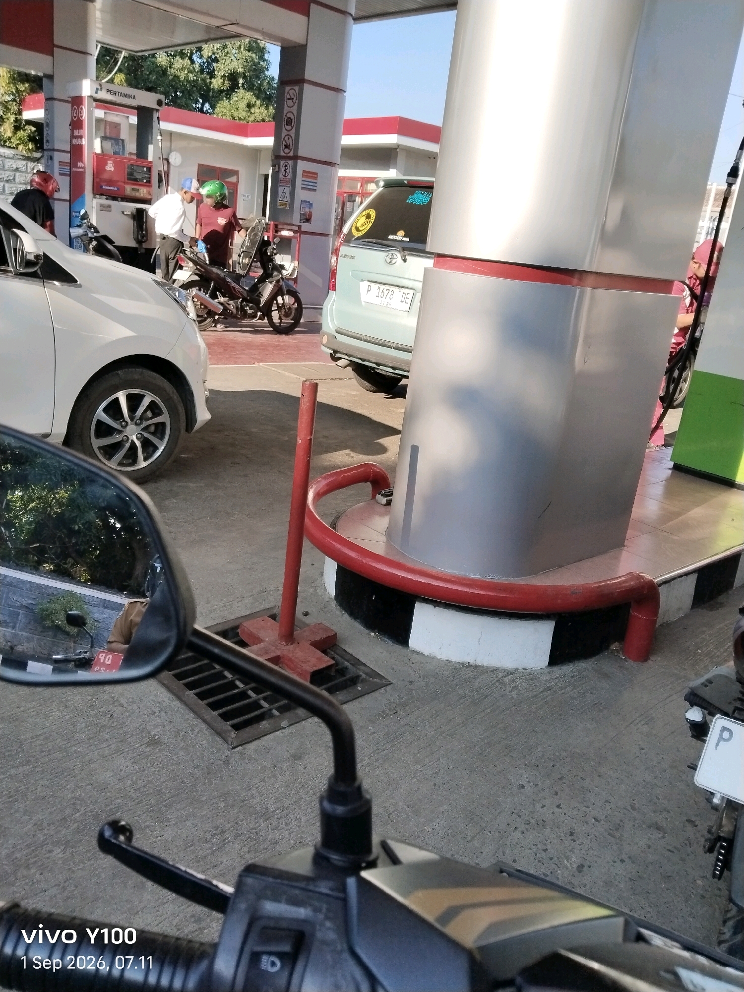 This image was taken at a Pertamina gas station in Situbondo, Indonesia. The scene depicts a white car and a motorcycle at a gas pump, with two individuals attending to the motorcycle. One person, wearing a white shirt and blue cap, appears to be interacting with the motorcycle's fuel tank. Another person, wearing a maroon shirt and green helmet, is standing next to the motorcycle. In the background, a silver van with a license plate ending in "DE" is visible. The ground is paved concrete, and a red metal pole with a red pipe barrier is present near the gas pump. The overall environment suggests a typical urban gas station setting under daylight.