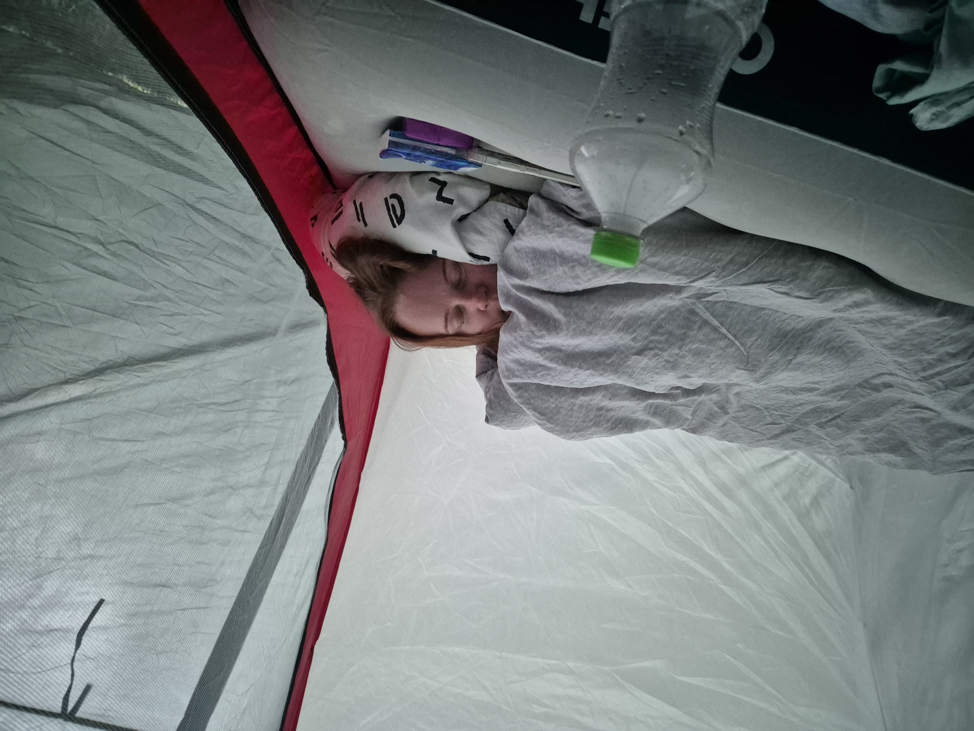 Inside a tent in Kehra, Estonia, a person is sleeping. The person is covered by a light gray blanket and is lying on an inflatable mattress. A water bottle with a green cap is visible in the foreground to the left of the person. To the right of the person, a small object with purple and blue colors is lying on the mattress. The tent has white and light gray fabric walls, with a red and gray trim visible on the right side.