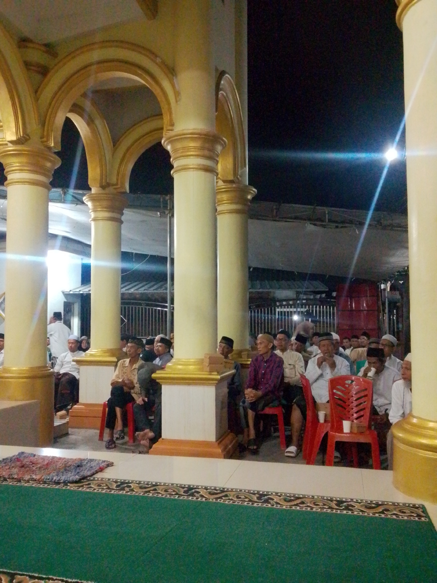 The image depicts a gathering of men, likely in a religious or community setting, situated under a covered outdoor area supported by ornate golden-trimmed pillars. The architecture suggests a mosque or a similar place of worship, possibly in Bandar Lampung, Indonesia, given the cultural attire and setting. The men are seated on chairs and on the floor, some are looking towards the camera, while others are engaged in conversation or listening. They are predominantly wearing traditional Indonesian clothing, including peci hats and sarongs. A prayer mat is visible on the floor in the foreground. The lighting is dim, indicating it is nighttime, with artificial lights casting a yellowish glow and creating lens flares. In the background, there appears to be a fenced area and some structures, suggesting an urban or semi-urban environment.