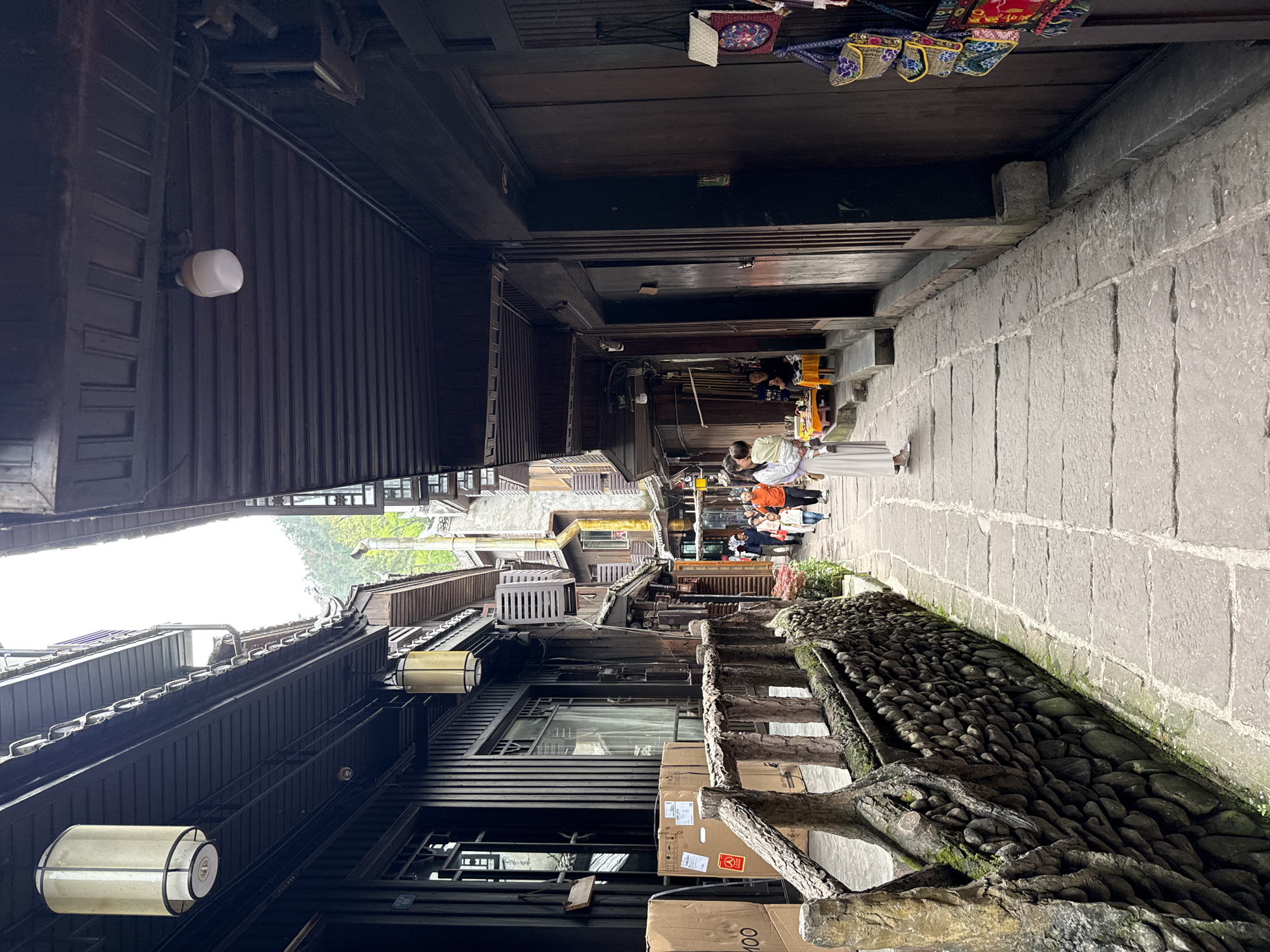 The image depicts a narrow alleyway in Fenghuang, China. The alley is paved with stone and lined with traditional wooden buildings. Several people are visible further down the alley, some appearing to be tourists. Along the right side is a stone wall or raised area with a collection of dark rocks. There are boxes near what may be a vendor's stall, adjacent to the stone feature. Lighting fixtures are attached to the buildings.