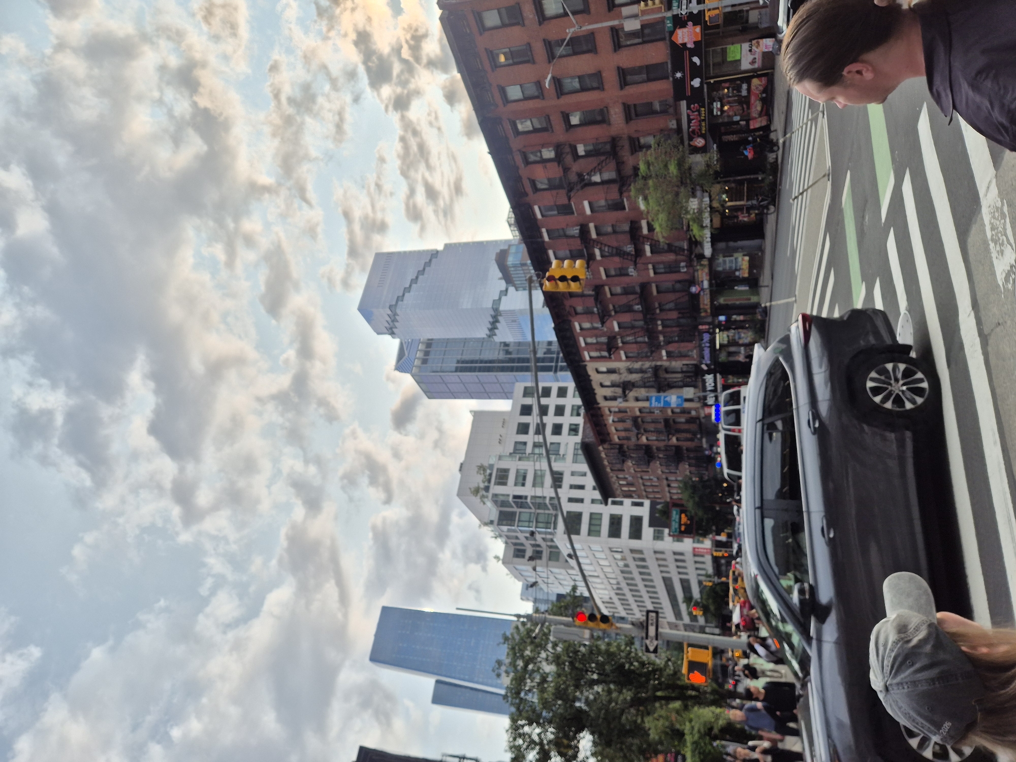 A street-level, eye-level, medium shot captures a bustling city scene in New York, USA. The foreground features two individuals on the sidewalk, one facing away from the camera, wearing a dark shirt and a baseball cap. Adjacent to them, a dark gray SUV is visible, partially obscuring the street. The street itself is marked with pedestrian crosswalks and painted green bike lanes. In the midground, a row of older, brick buildings with fire escapes lines the street, housing various businesses. Beyond these, a cluster of modern, glass skyscrapers dominates the skyline, their reflective surfaces creating a striking contrast with the older architecture. A traffic light displaying red is prominent on the left side of the frame, indicating a stop. The sky above is filled with a dramatic, cloudy expanse, suggesting either early morning or late afternoon light. The scene appears to be in a commercial and residential area of the city, likely in Manhattan.