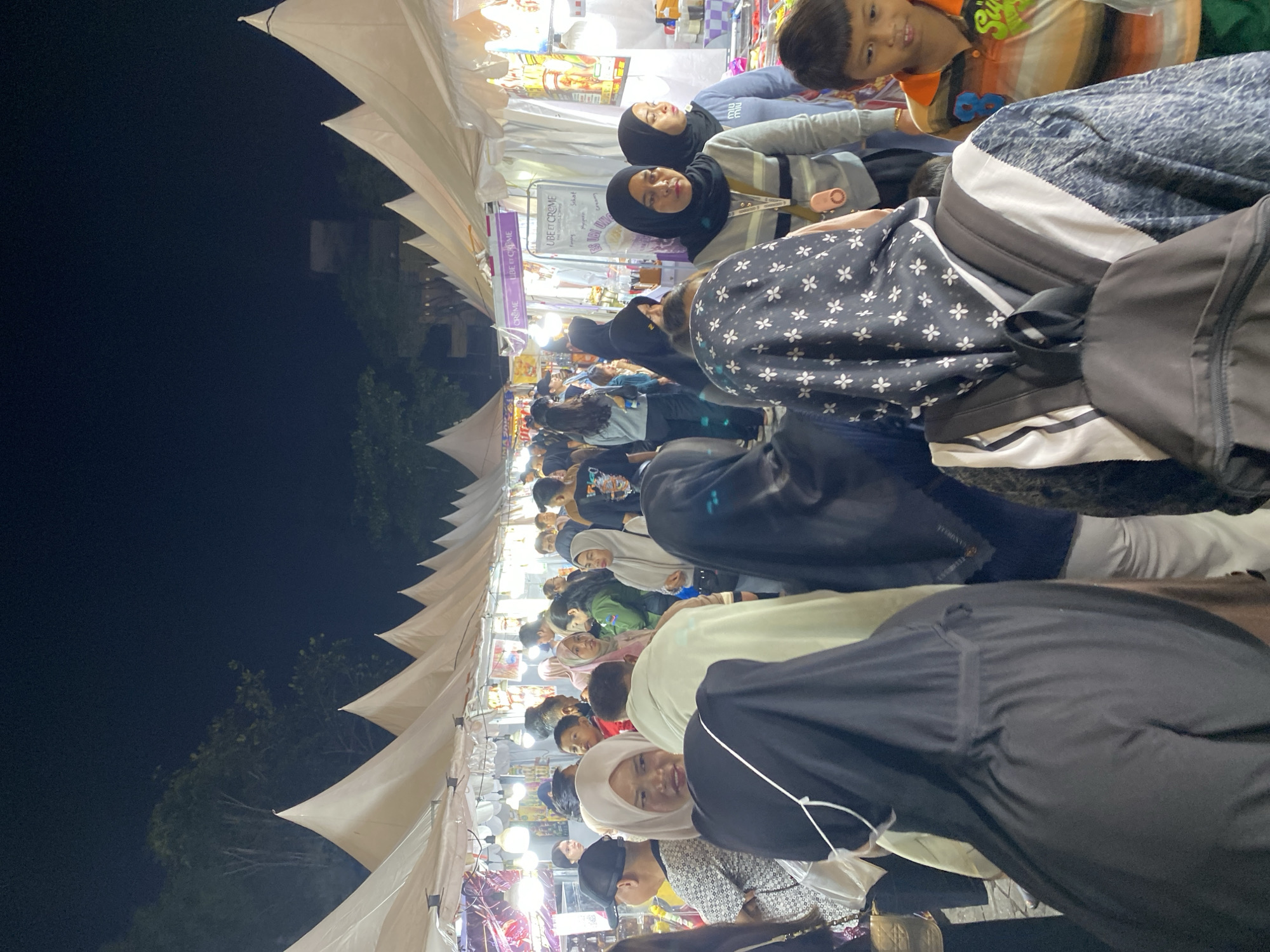 The image depicts a bustling night market scene in Banjarmasin, Indonesia, likely near a waterfront or a popular public area given the presence of numerous stalls and crowds. The atmosphere is lively, with many people browsing and interacting. The stalls are covered by white, triangular tents, illuminated by strings of lights, creating a warm and inviting ambiance.
The foreground features several individuals, including a woman wearing a black hijab and a patterned headscarf, with a backpack on her back. To her left, another person in a black abaya is partially visible. In the mid-ground, a crowd of people of various ages are visible, many wearing hijabs and traditional Indonesian attire. The people are dressed in casual clothing suitable for an outdoor evening event. Some individuals appear to be vendors, tending to their stalls, while others are shoppers exploring the offerings.
The stalls themselves display a variety of goods, although the specific items are difficult to discern due to the lighting and distance. One stall in the background has a sign that reads "UBE ET CREME," suggesting food or beverage items. Other stalls appear to sell clothing, accessories, or souvenirs. The overall environment is typical of a vibrant Asian night market, characterized by its energetic atmosphere, diverse crowd, and variety of retail options. The backdrop includes trees and some building structures, indicating an urban setting within Banjarmasin. The darkness of the night sky contrasts with the bright lights of the market, highlighting the lively activity below.