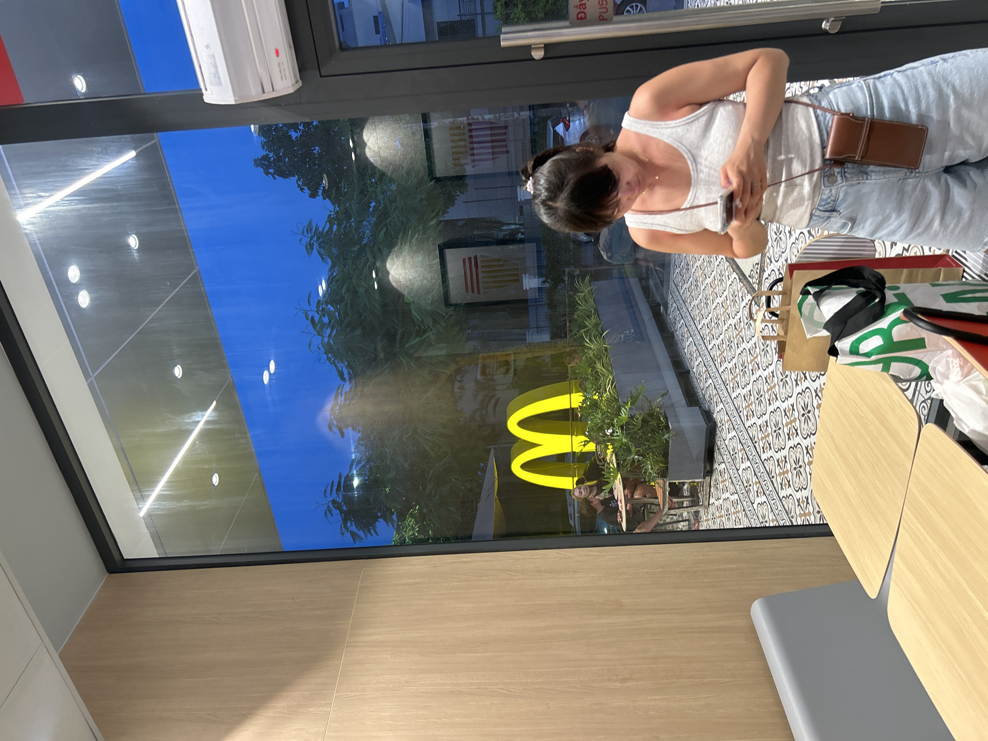 A woman stands looking down at her phone in a McDonald's restaurant in Nha Trang Ward, Vietnam. She is wearing a white tank top and denim shorts, with a small brown purse slung over her shoulder. The McDonald's logo is visible through the large window behind her, along with trees and the night sky. The interior of the restaurant features light wood paneling, gray seating, and wooden tables. The floor is tiled with a patterned design. Two shopping bags, one brown and one white with green lettering, are on a table to her left. In the background, through the window, two people are seated at a table outside.