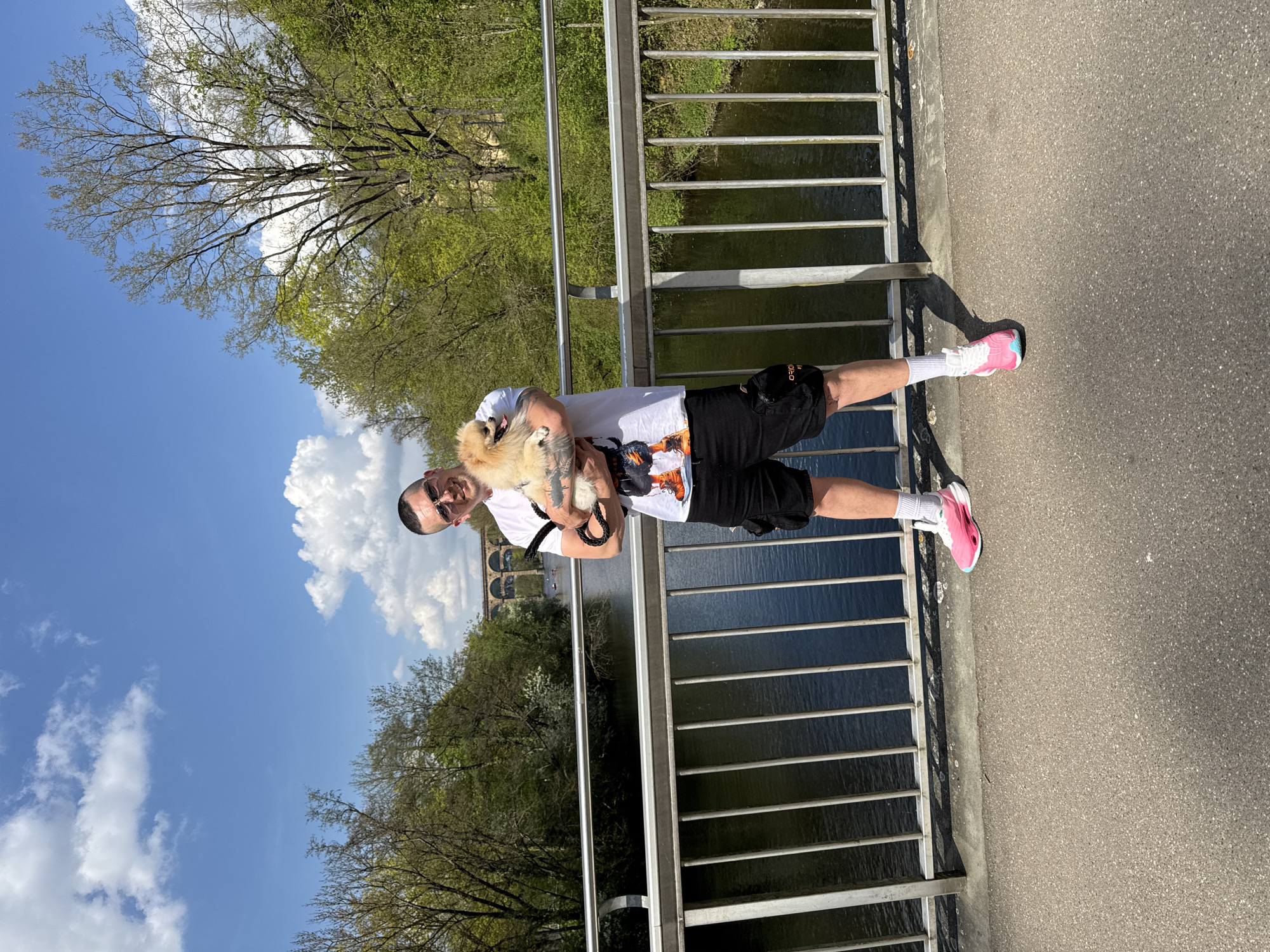 A man holds a small, fluffy, light-colored dog on what appears to be a bridge or pedestrian walkway. He is facing the camera, wearing sunglasses, a white t-shirt with a graphic print, black shorts with a partially visible "GRON" like text, white socks, and distinctive pink and light blue athletic shoes. Tattoos are visible on his arms, and he has a slight smile. The small dog, possibly a Pomeranian, is held in his arms, looking forward.
The scene unfolds on a sunny day, with bright daylight casting clear shadows. A sturdy metal railing separates the paved walkway from a tranquil body of water below, likely a river or canal, which reflects the clear blue sky adorned with white, fluffy clouds. On the far side of the water, lush green trees indicate a vibrant natural environment. Prominently in the background stands a large, multi-arched historical viaduct, characteristic of the Bietigheim Viaduct over the Enz river in Bietigheim-Bissingen, Germany, which serves as a key location cue. The man and dog appear to be enjoying a leisurely moment outdoors.